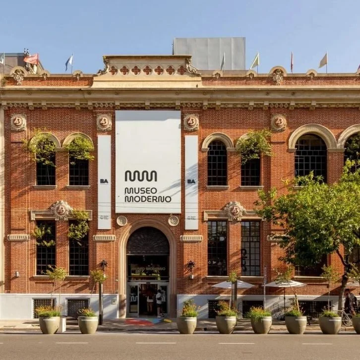 Front view of a historic red brick building with arched windows and decorative details, housing the Museo Moderno, with potted plants and trees outside.