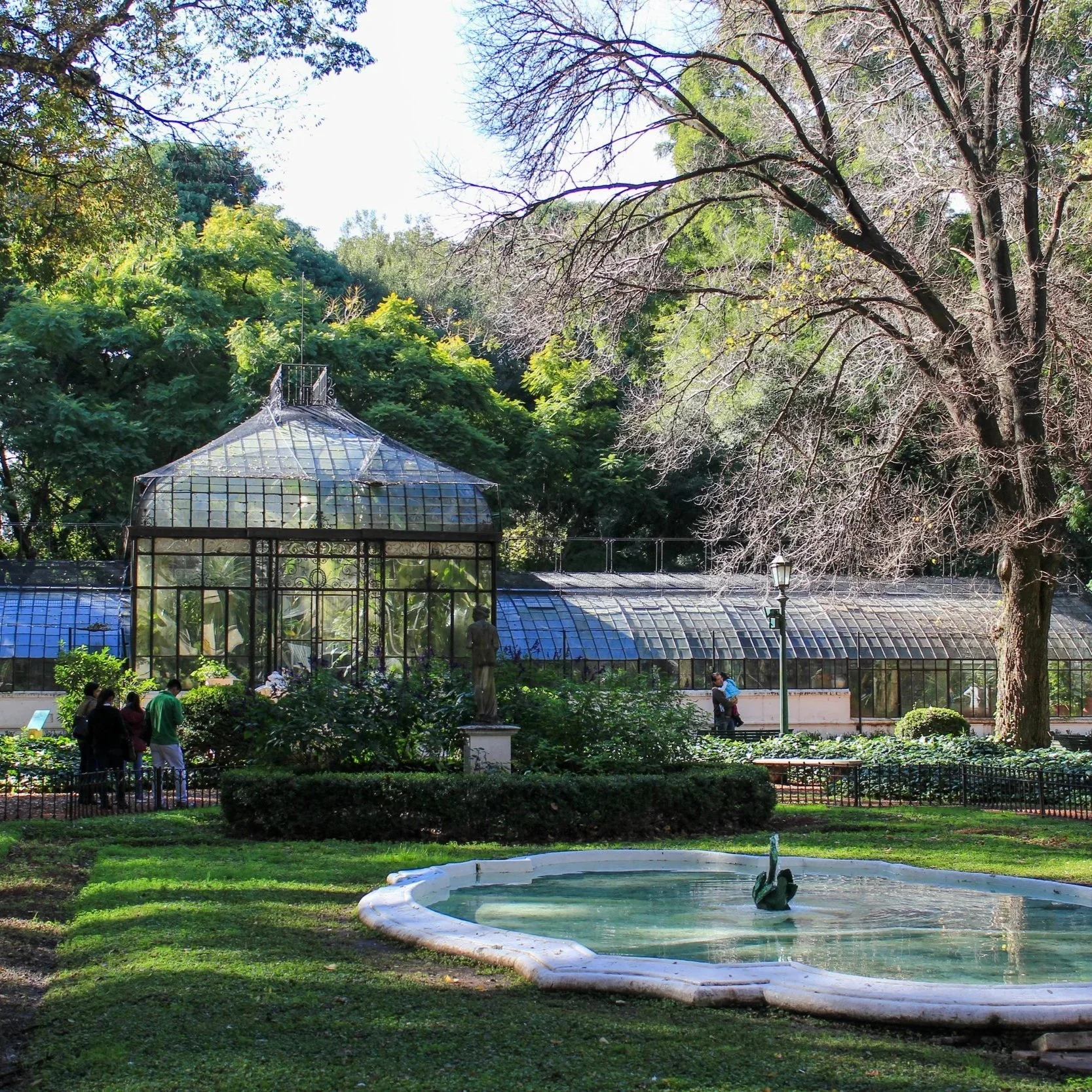 A garden with a small pond and fountain in the foreground, surrounded by greenery and trees. A glass greenhouse is visible in the middle distance, with several people walking or standing nearby. The scene is bathed in sunlight with blue sky overhead.