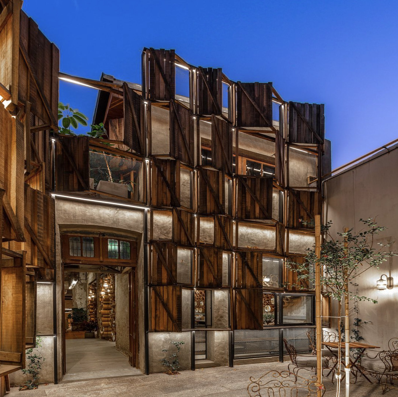 Night view of a multi-story building with a wooden and glass exterior, illuminated by outdoor lighting, with potted plants and a small garden area outside.