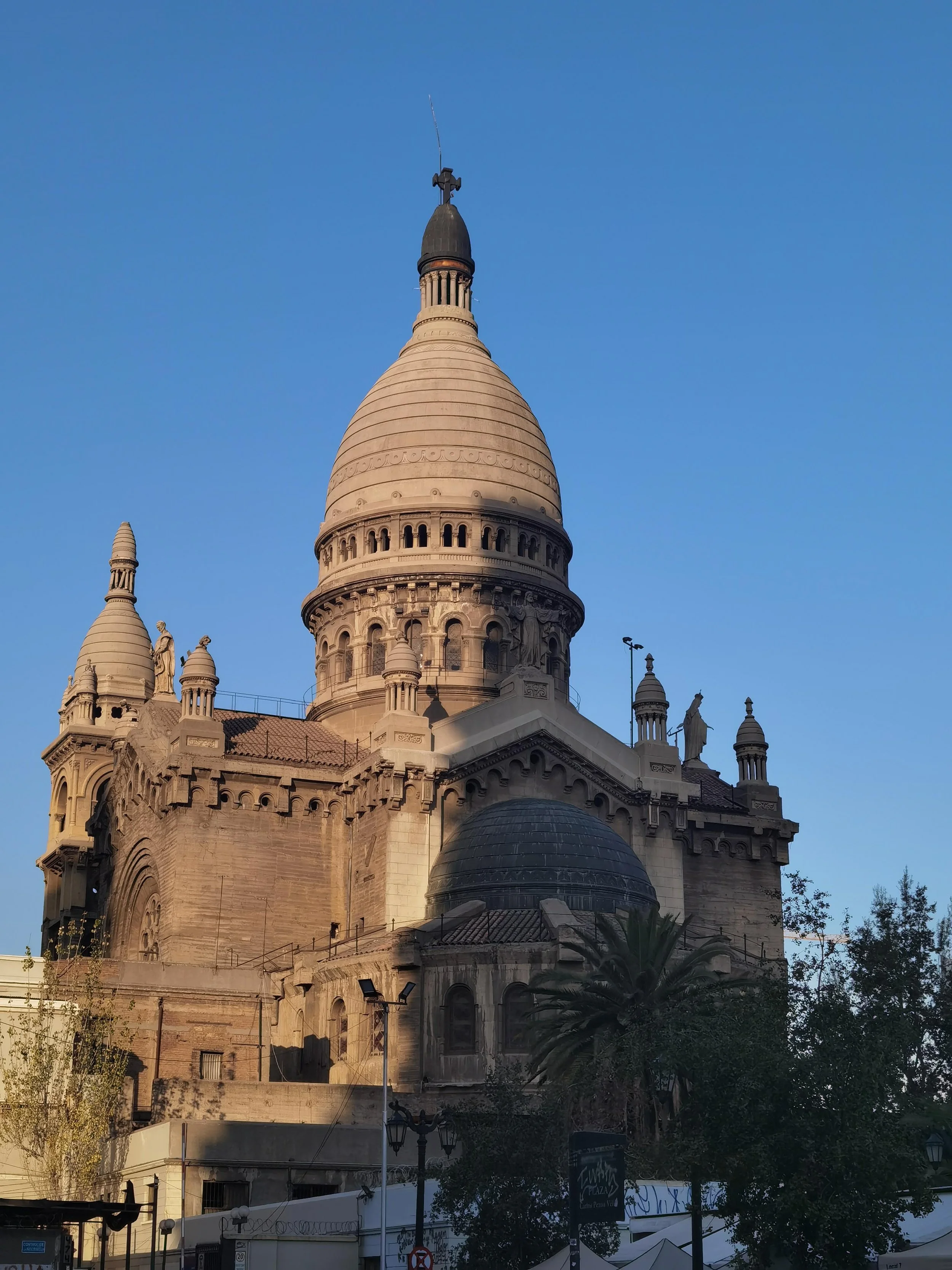 The image features a historic stone church or cathedral with intricate architectural details, several domes, turrets, and statues, set against a clear blue sky, with nearby trees and modern structures in the foreground.