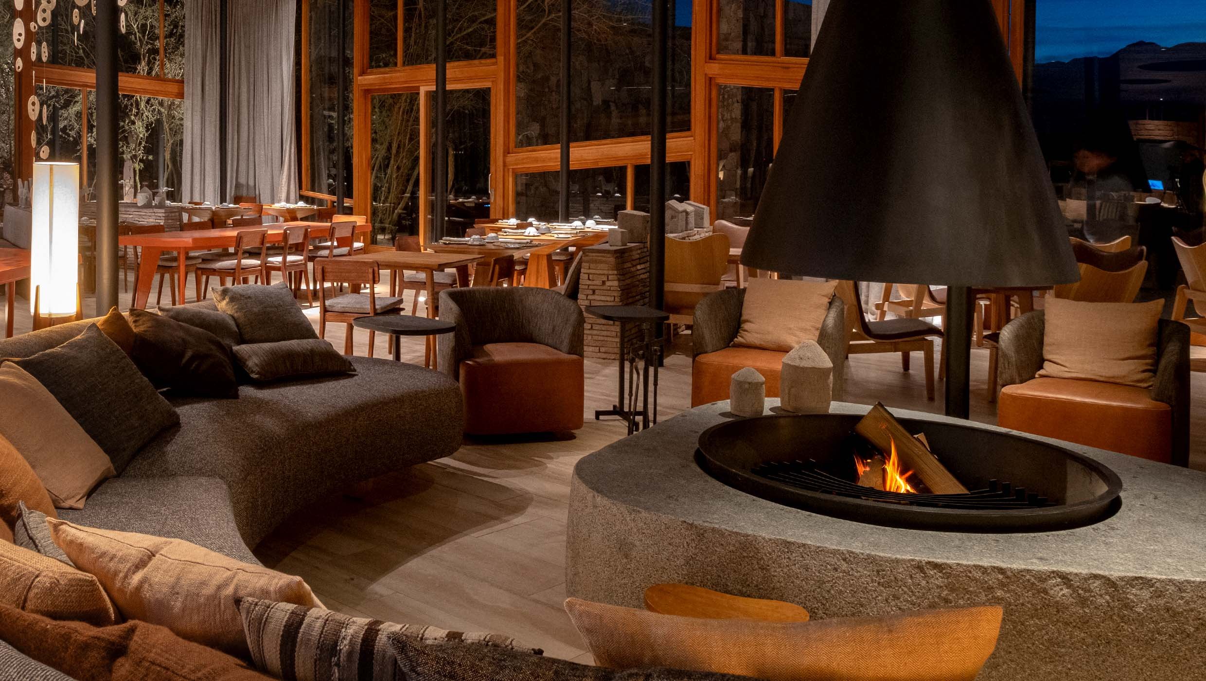The warm timber lounge and dining space of a design led Atacama lodge, with a central fire and floor to ceiling windows onto the desert night