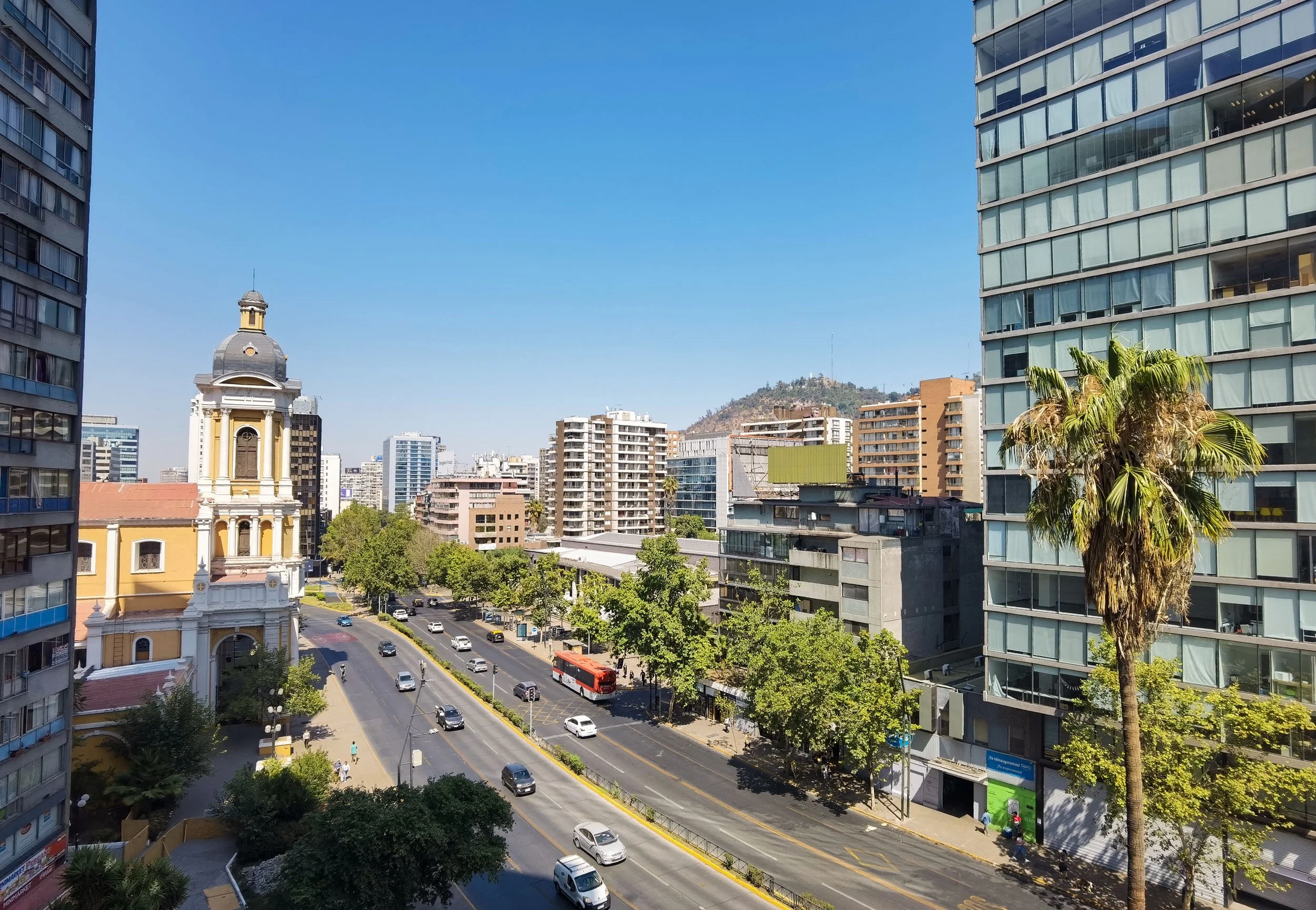 Cityscape with high-rise buildings, a church with a yellow facade and clock tower, vehicles on the street, and a hill with communication antennas in the background under a clear blue sky.