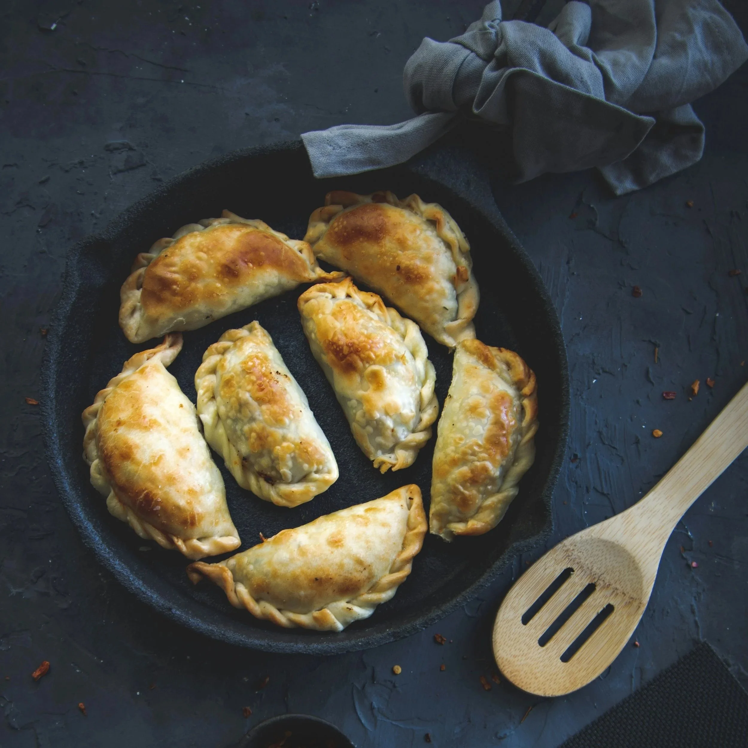 Fried dumplings in a black skillet, with a wooden spatula nearby, on a dark textured surface.