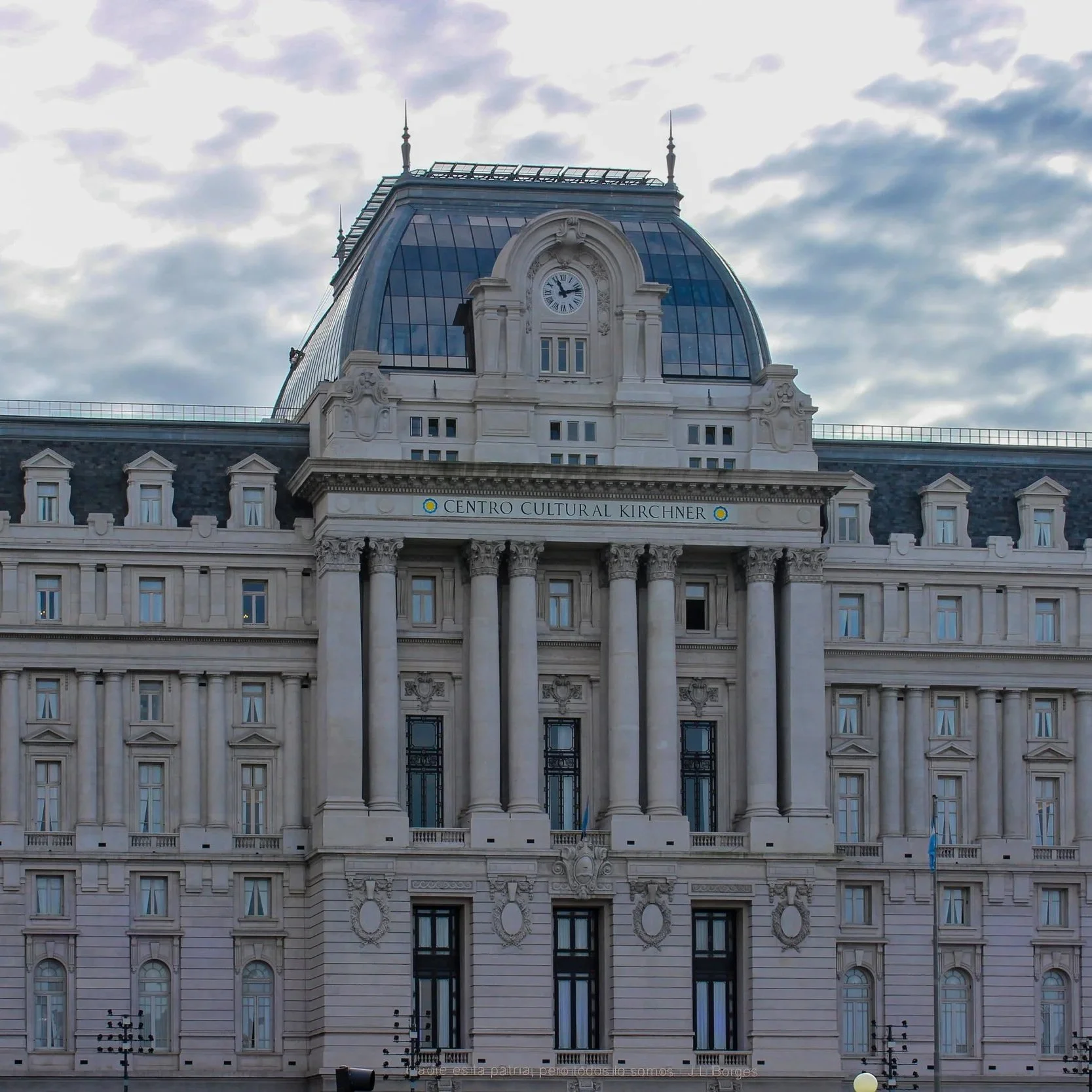 A historic building with a grand classical façade, columns, and decorative architectural details, labeled 'Centro Cultural Kirchner' at the entrance. The building has a prominent clock and a glass dome on top, with a cloudy sky in the background.