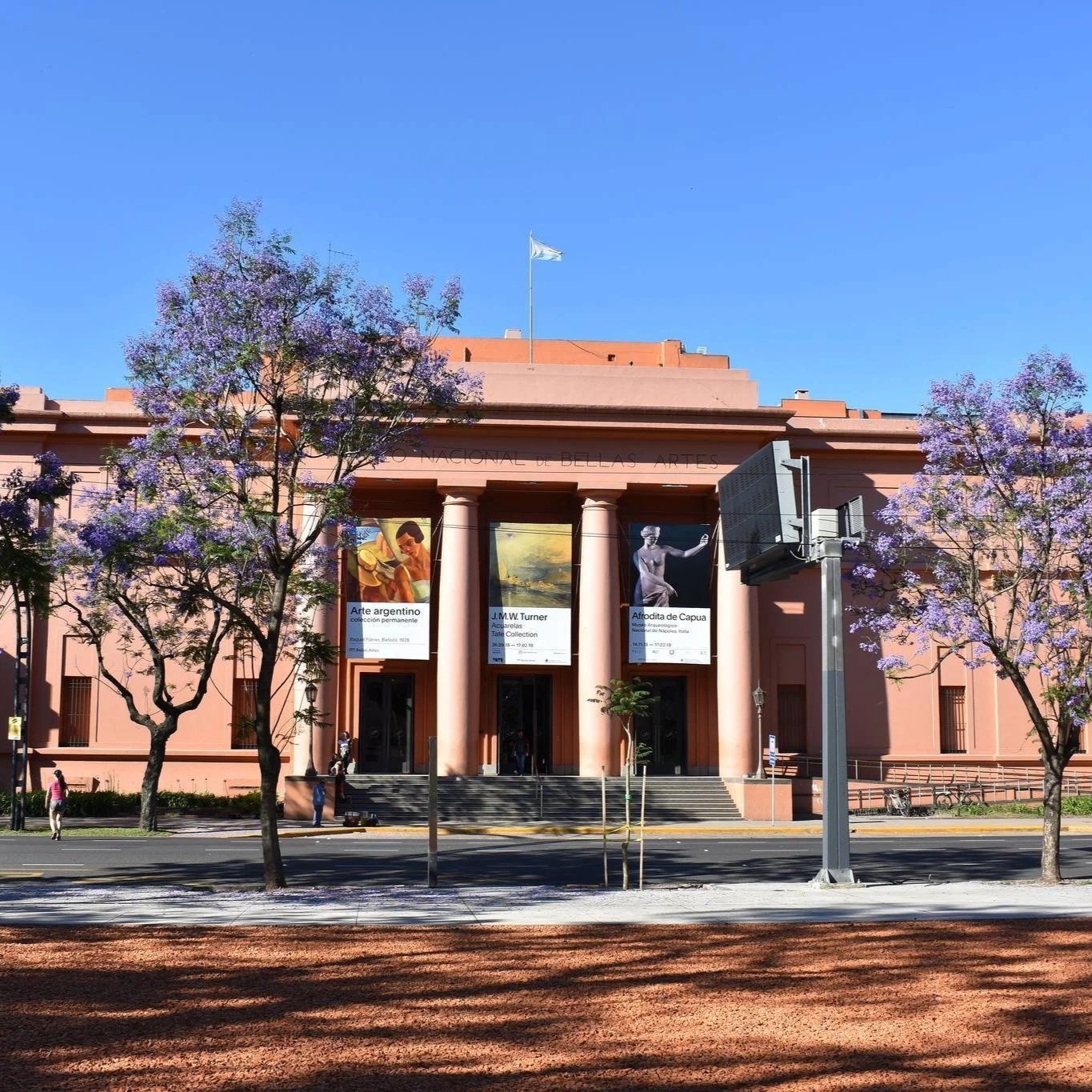 Pink neoclassical building with columns and banners displaying art exhibitions, located on a city street with purple flowering trees and a blue sky.