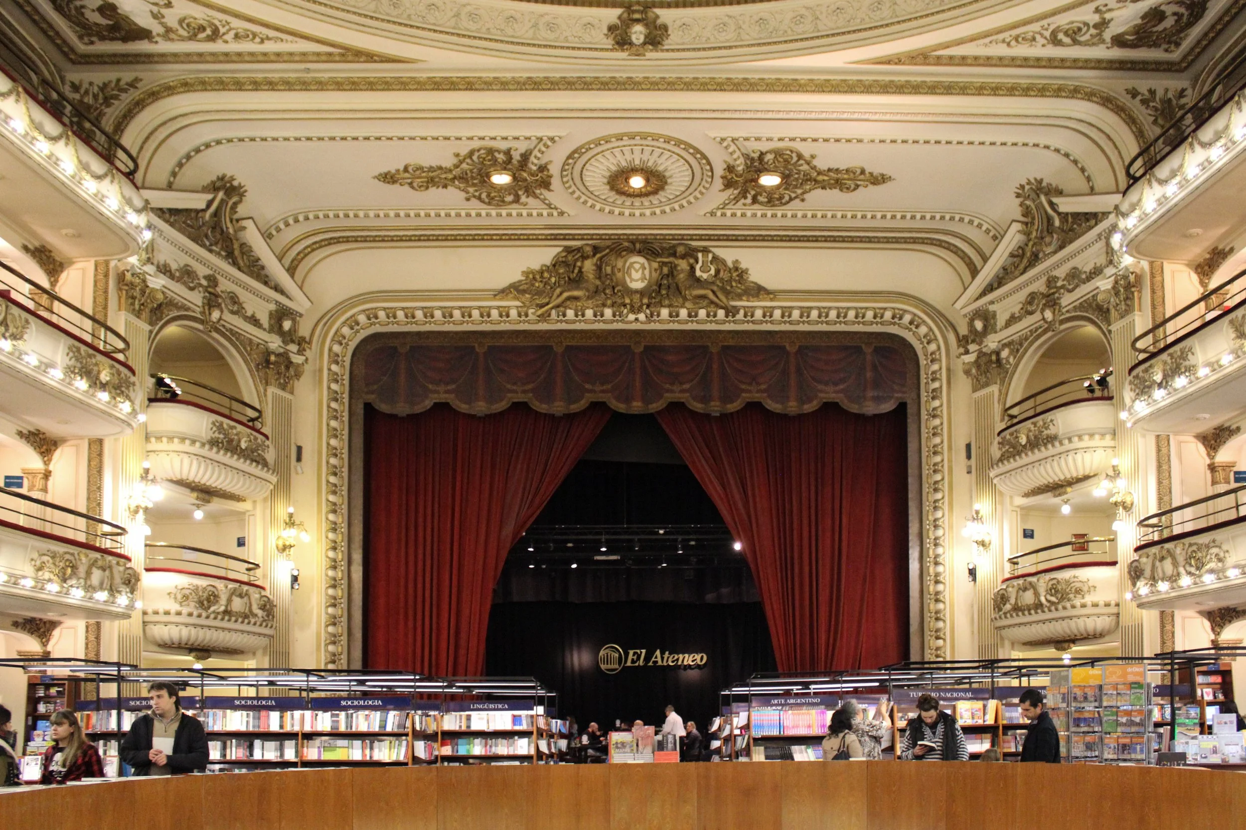 Interior of a grand, ornate theater with a stage and red curtain, balconies with decorative railings, and people browsing bookshelves in front.