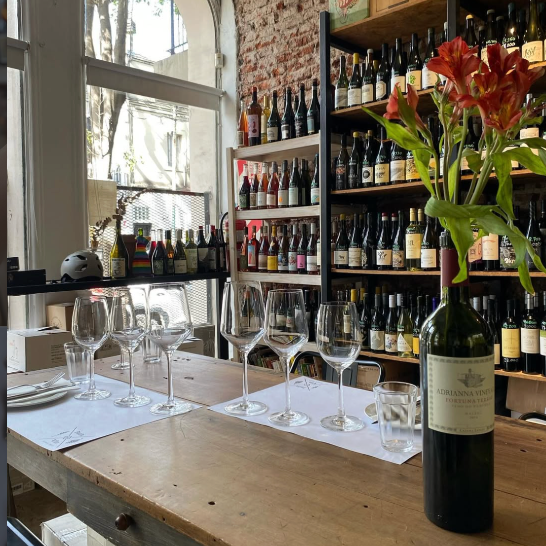 A wine tasting setup with multiple empty wine glasses on a wooden table, a bottle of red wine with a flower in it, and shelves filled with various bottles of wine in a well-lit room with large windows and a brick wall.