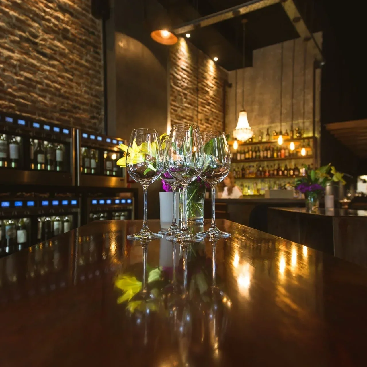 Four empty wine glasses on a polished wooden table in a dimly lit restaurant with exposed brick walls, bottles on shelves, and flowers in vases.