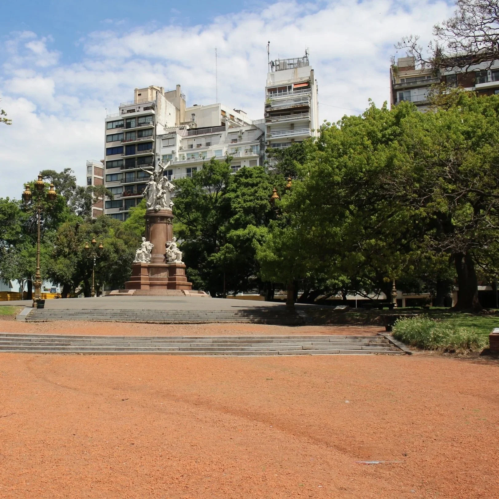 A park with a statue in the center, surrounded by green trees and street lamps. In the background, there are tall residential buildings with balconies, set against a partly cloudy sky.