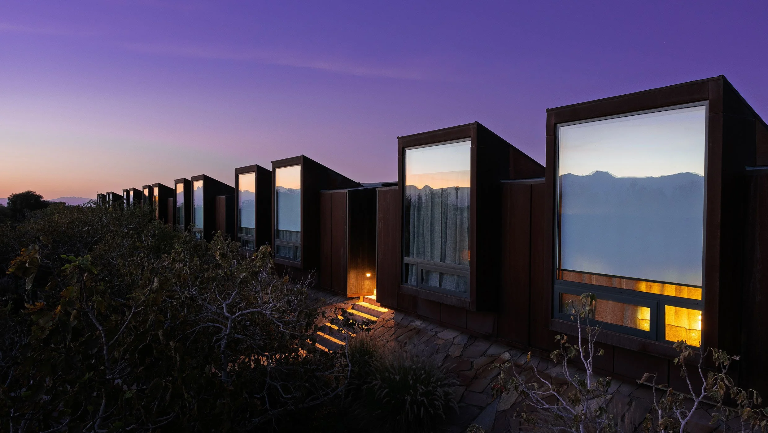 A low timber desert lodge glowing at twilight beneath a violet sky, with the Andes silhouetted along the horizon of the Atacama