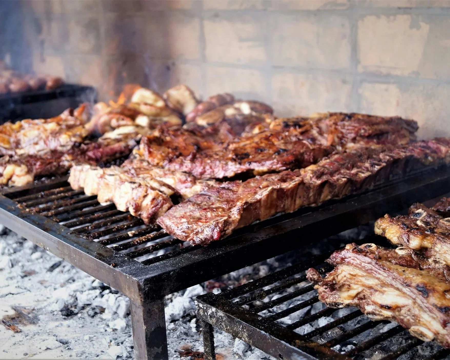Barbecue meat grilling on a commercial grill over hot coals with a brick wall background.