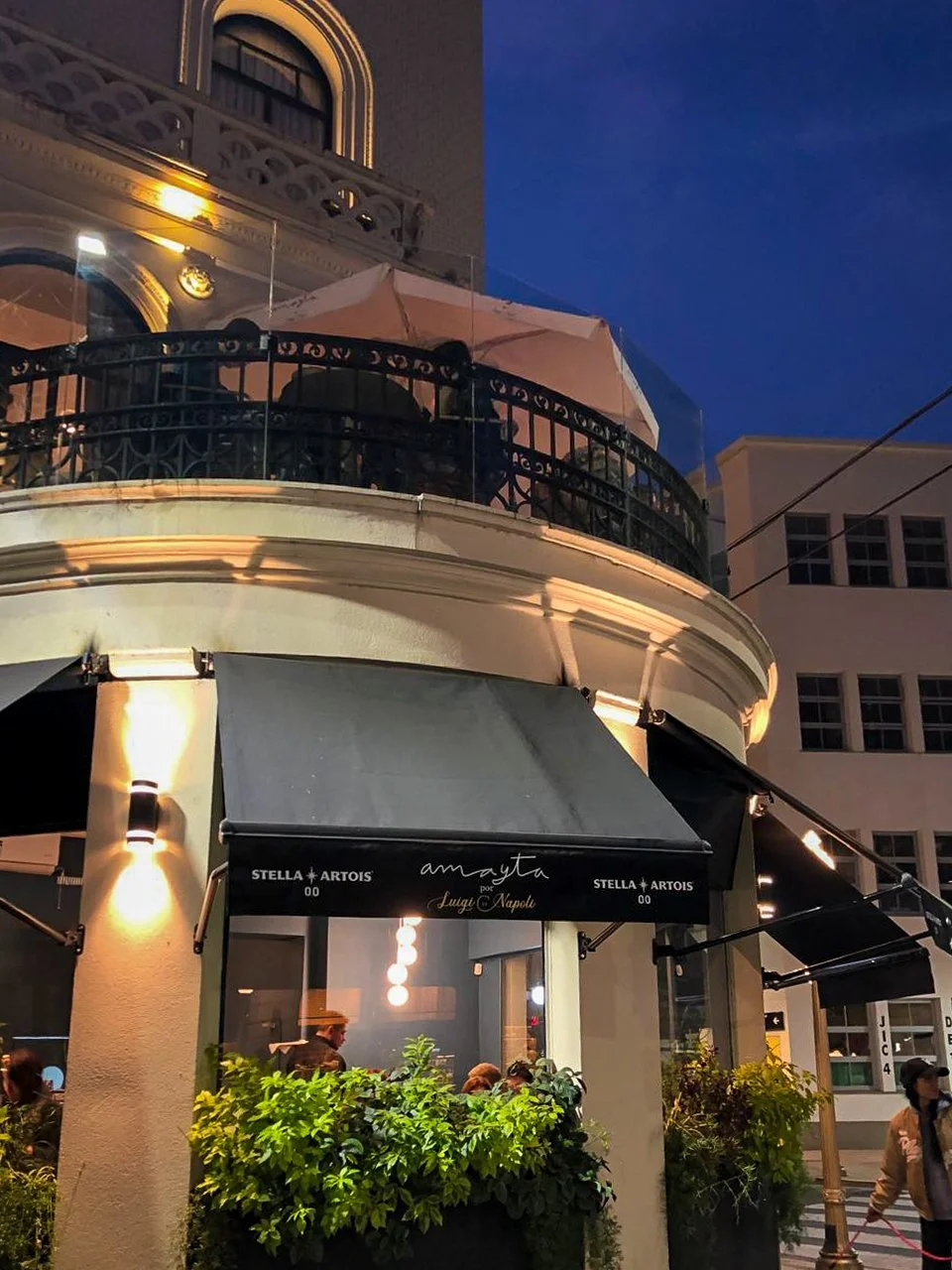 Night view of a restaurant named Amayta with outdoor seating, located on a corner of a building with a balcony and decorative railing, illuminated by street and building lights.
