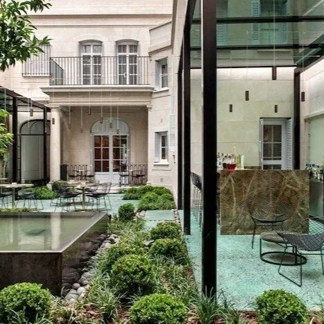 A view of a modern outdoor courtyard with a small pool, greenery, and outdoor seating, surrounded by a glass wall and an elegant building with balconies.