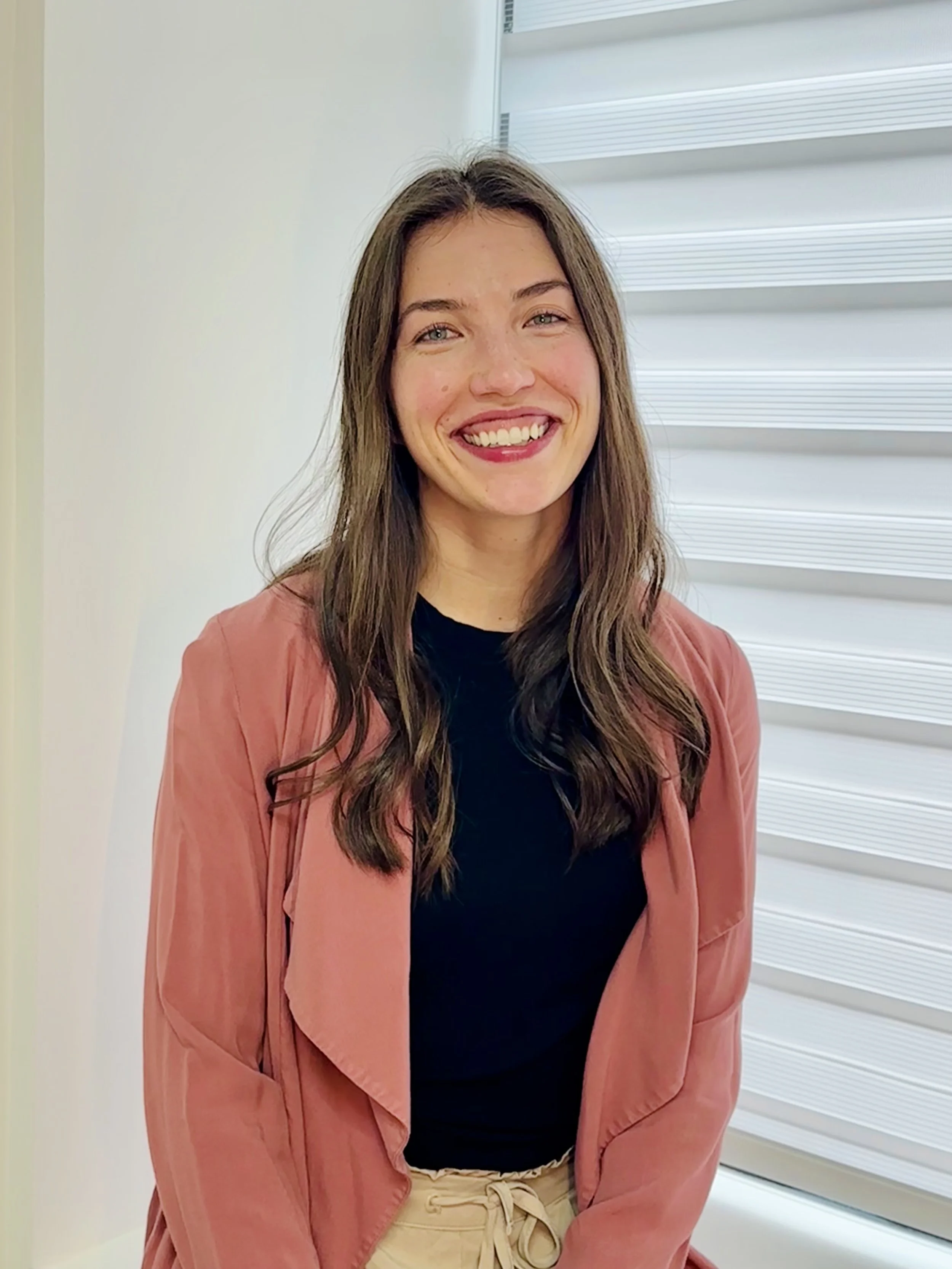A woman with long brown hair, smiling, wearing a black top and tan pants, standing in front of white horizontal blinds.
