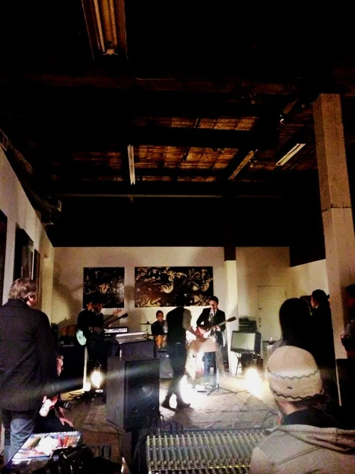 A live band performing on stage in a cozy indoor venue with wooden ceiling and abstract art on the wall, and audience members watching.