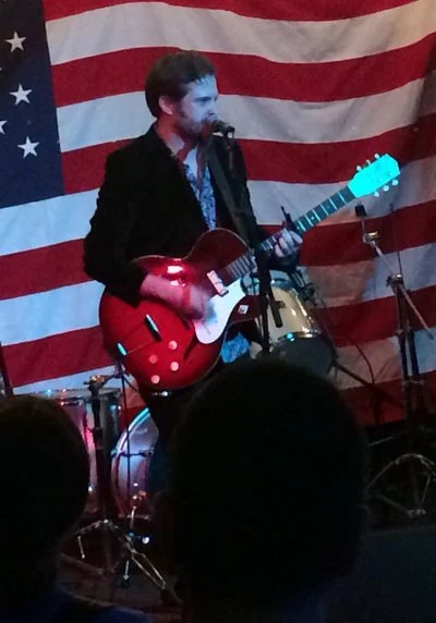 Man playing a 1966 Harmony Rocket guitar on stage in front of an American flag