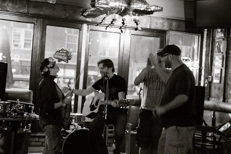 Man with guitar surrounded by three people clapping and laughing