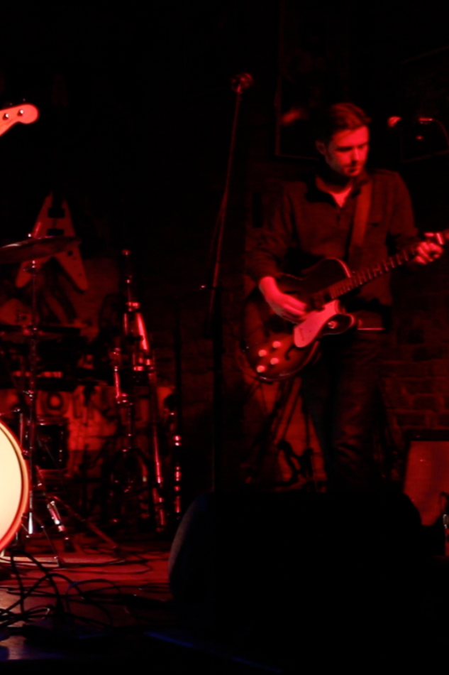 A musician playing an electric guitar on stage under red lighting, with drums visible in the background.