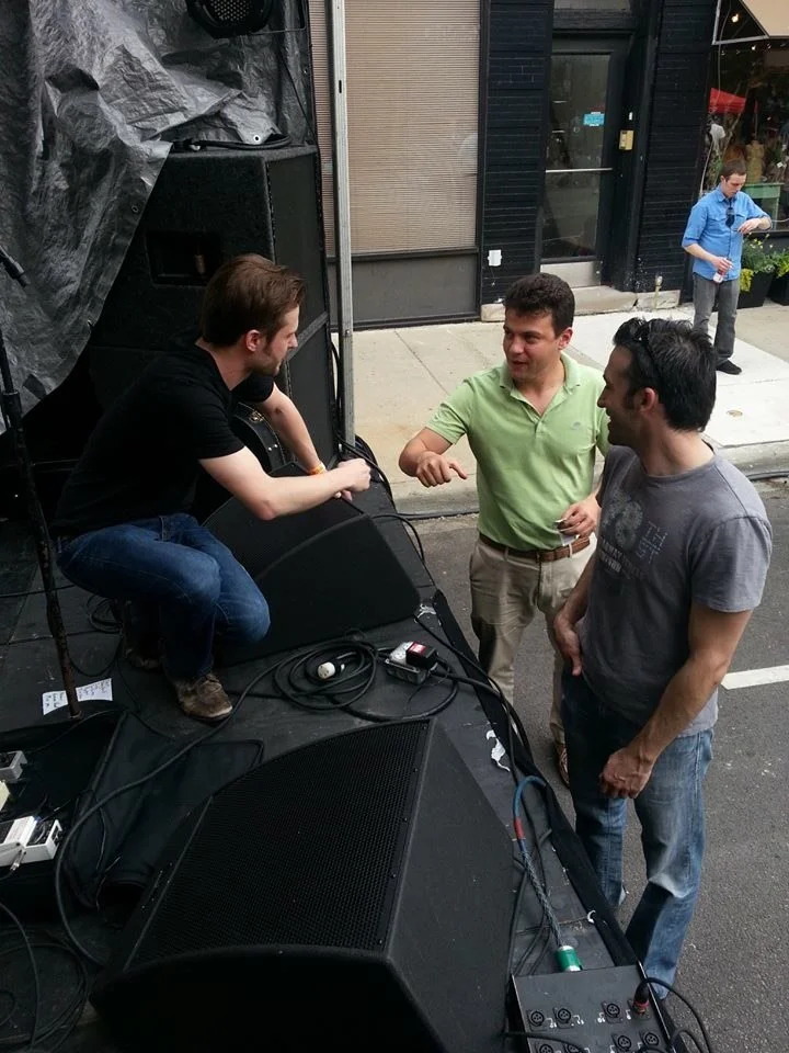 Three men engaged in conversation near sound equipment outdoors, with two of them standing and one sitting on the equipment.
