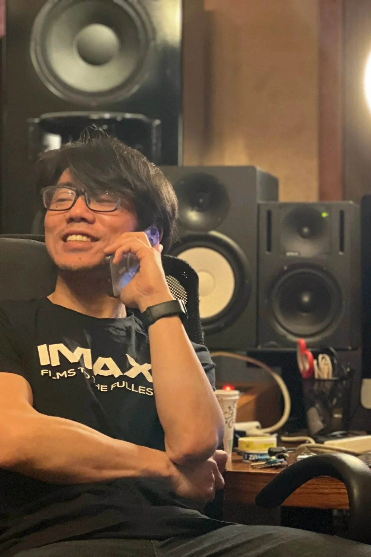 A man with black hair, glasses, and a black IMAX t-shirt sitting in a recording studio, smiling, holding a phone to his ear, with large black and white speakers and audio equipment in the background.