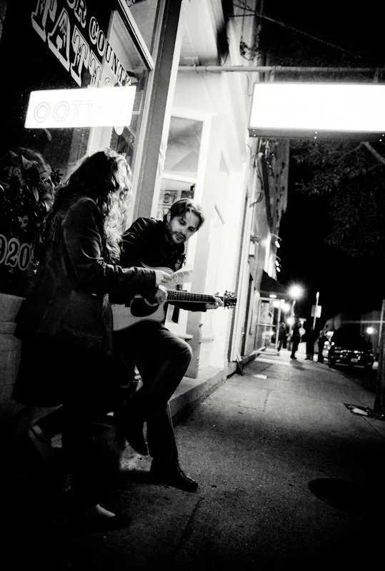 Woman holding paper next to man with guitar. They are standing on the street outside a music venue