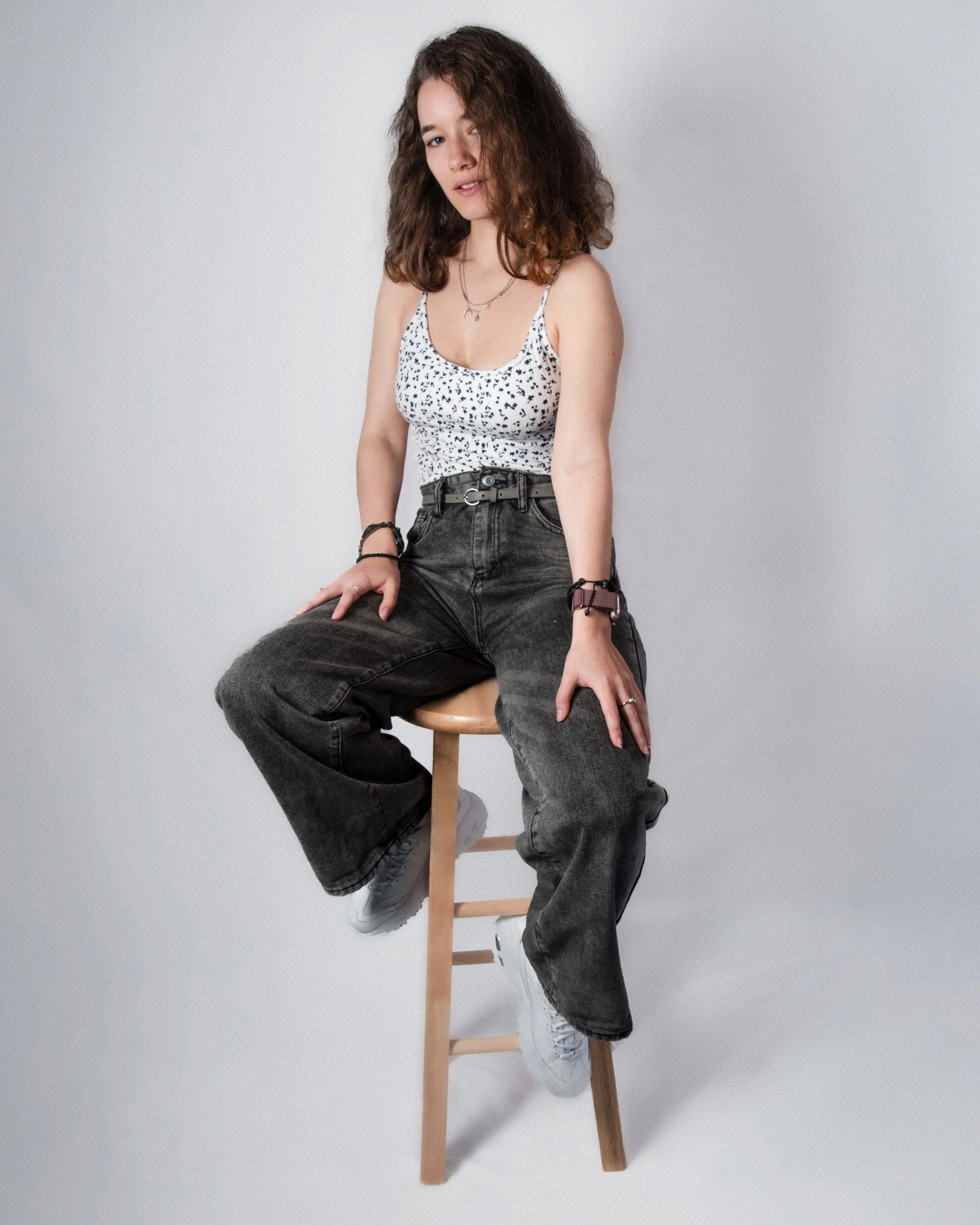 A young woman with curly brown hair sitting on a wooden stool against a plain background, wearing a white tank top with black pattern, black jeans, and white sneakers.