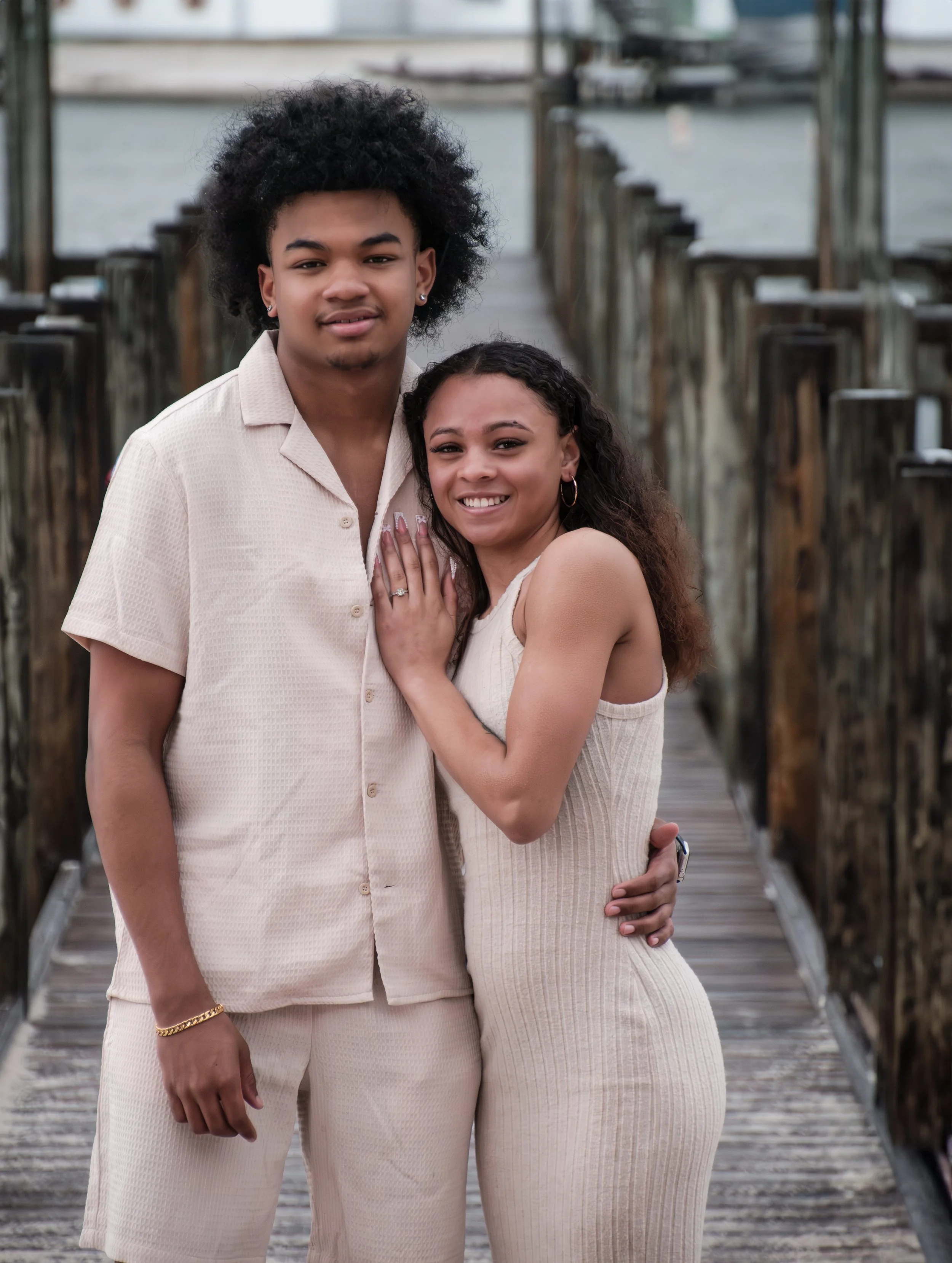 A smiling couple standing on a wooden pier near the water, with the man having curly black hair and the woman with long, curly dark hair.