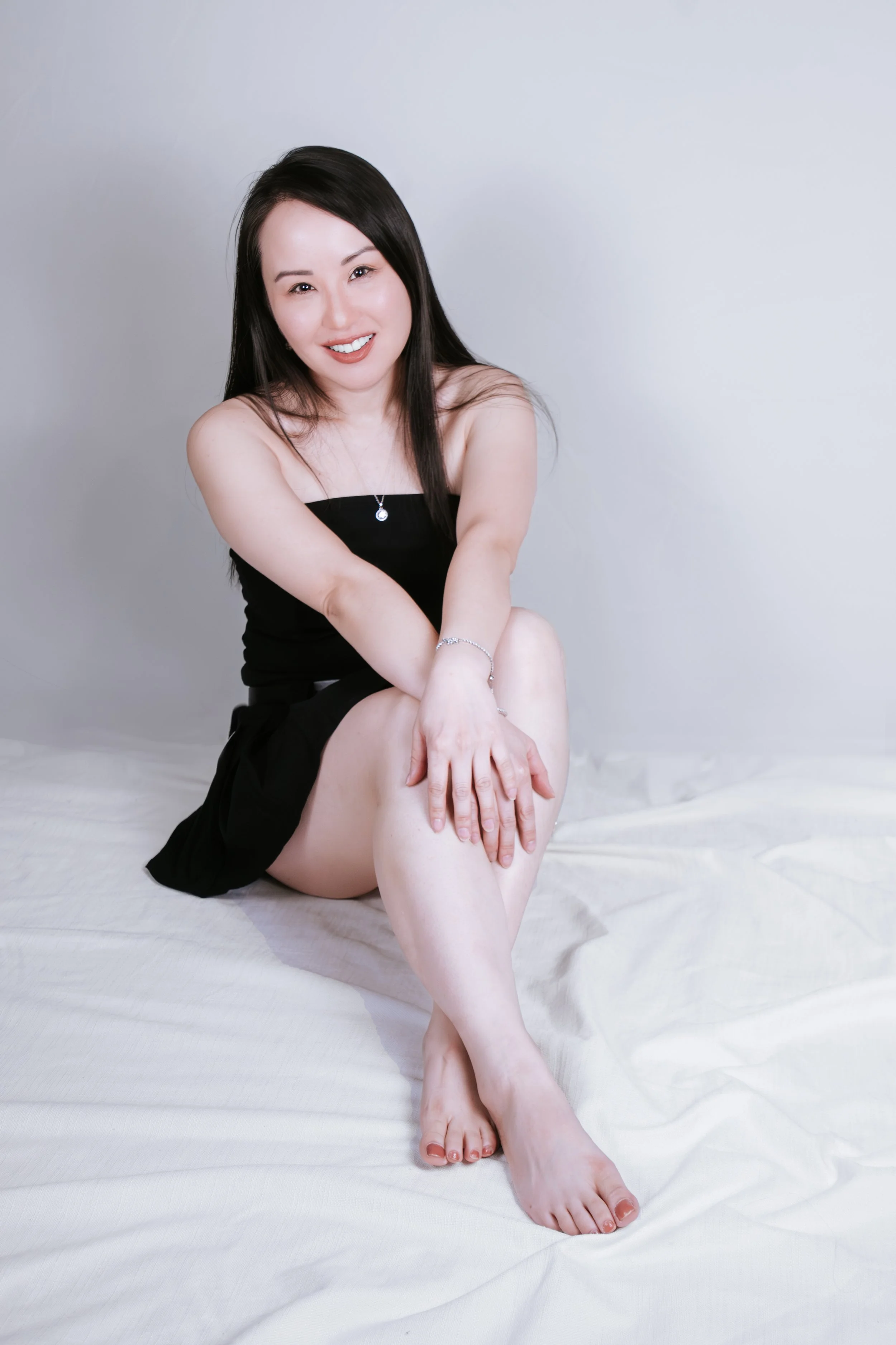A woman with long black hair sitting on a white bed, wearing a black strapless dress, smiling and looking at the camera.