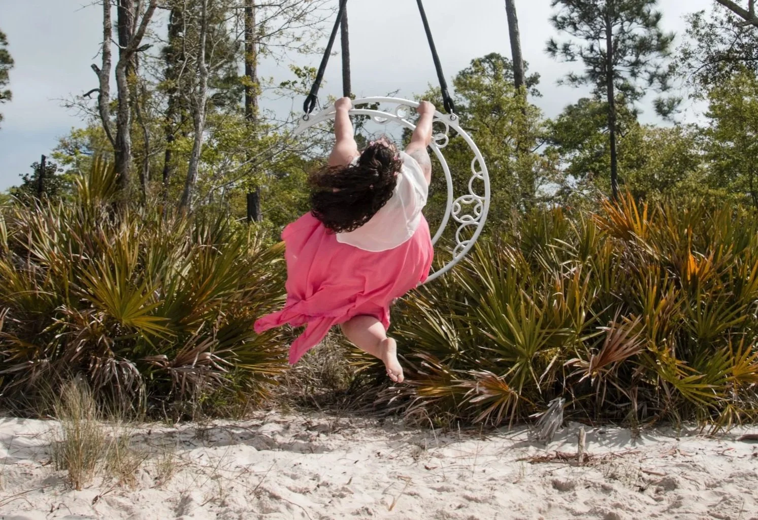 A person hanging upside down from a horizontal bar in a wooded outdoor area, using a red and a blue pole for support.