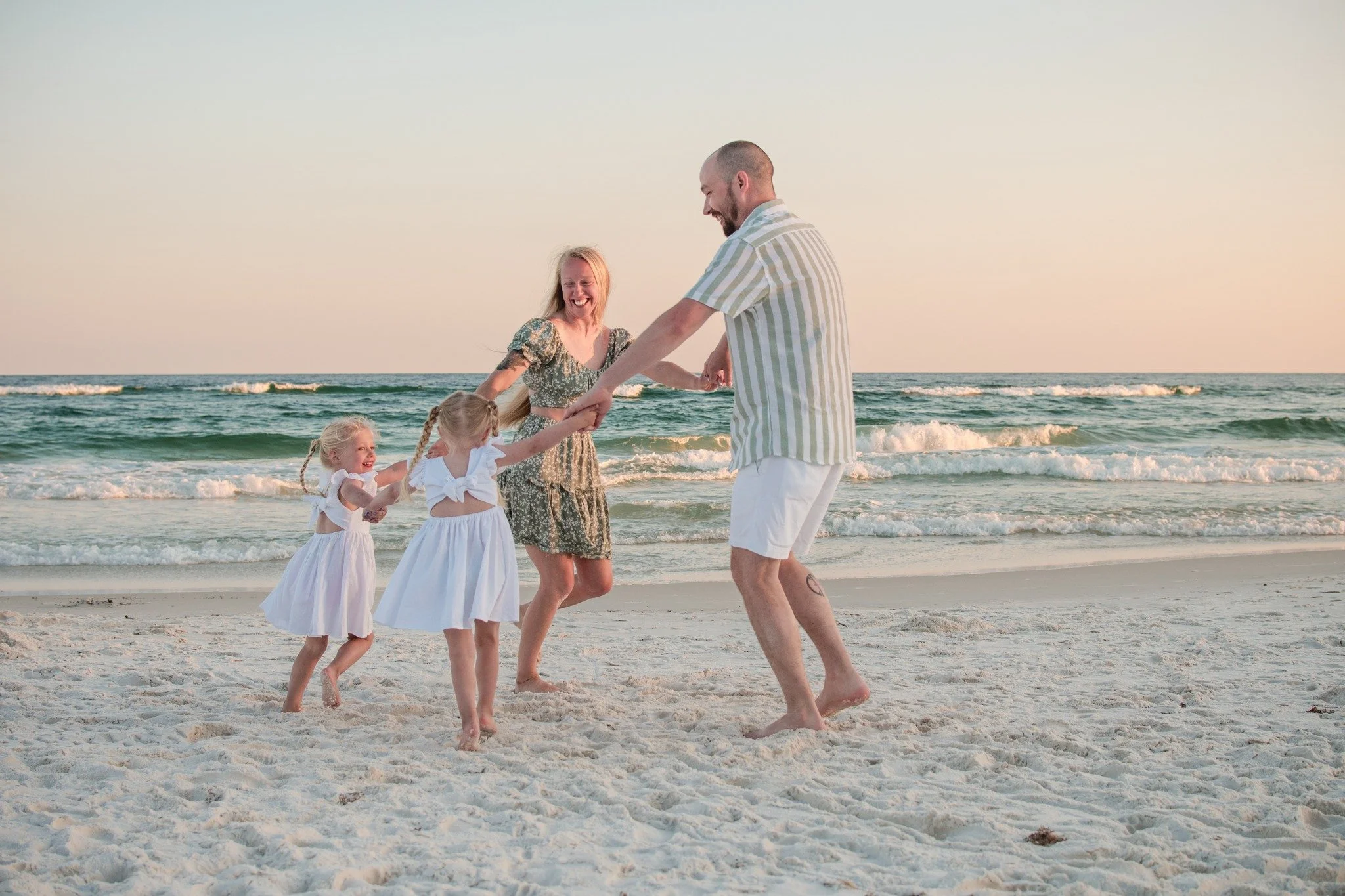 A family of four, including two young girls, two adults, are playing and dancing on the beach at sunset, holding hands and smiling.