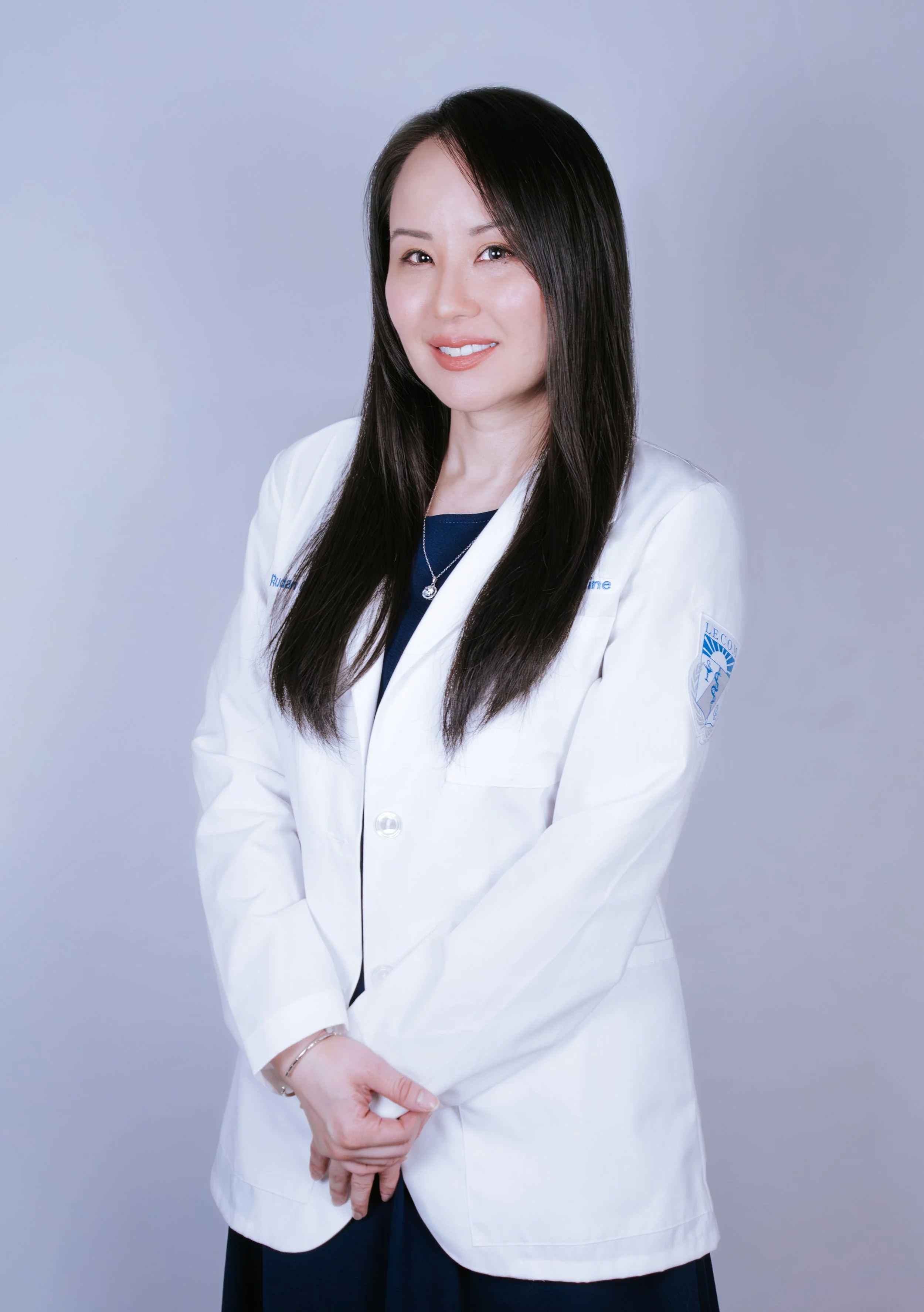 A woman with long dark hair wearing a white medical coat and a necklace, standing against a light background.