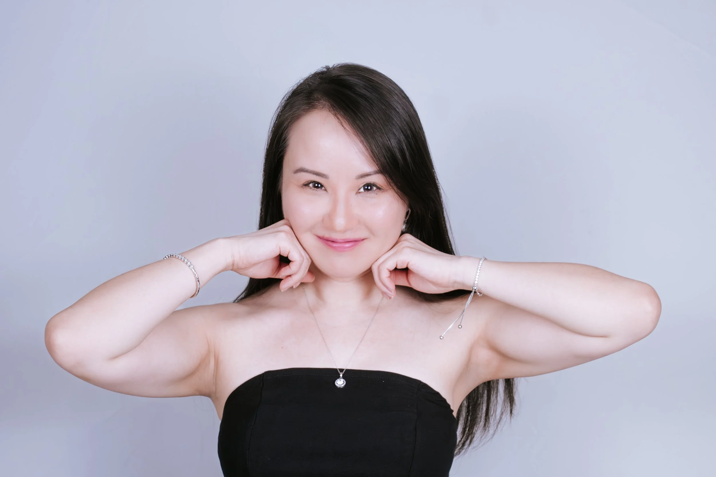 A woman with long dark hair smiling and holding her face with both hands, wearing a black strapless top and jewelry, against a plain light-colored background.