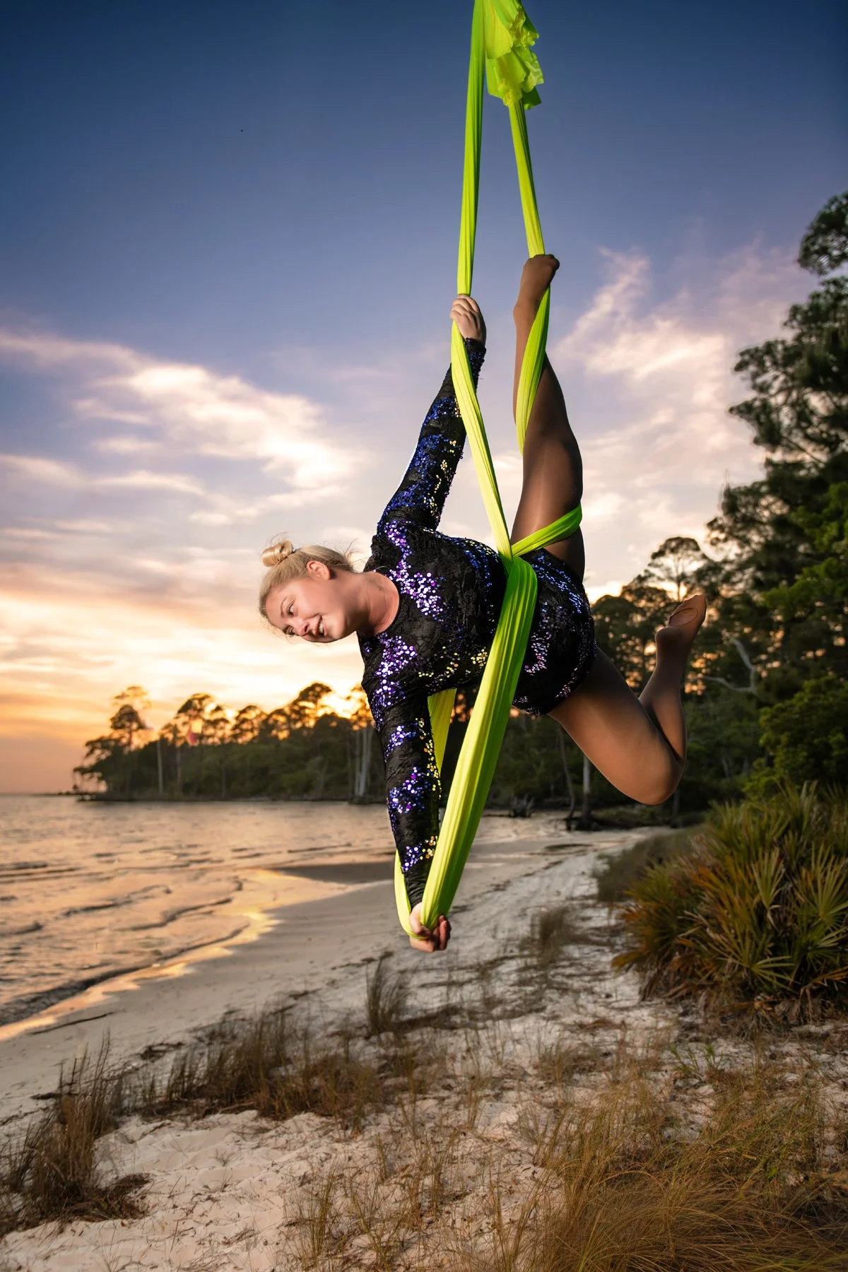 A woman practicing aerial silks outdoors among trees, hanging upside down with her right hand reaching towards her left foot while holding blue aerial silk with her left hand.
