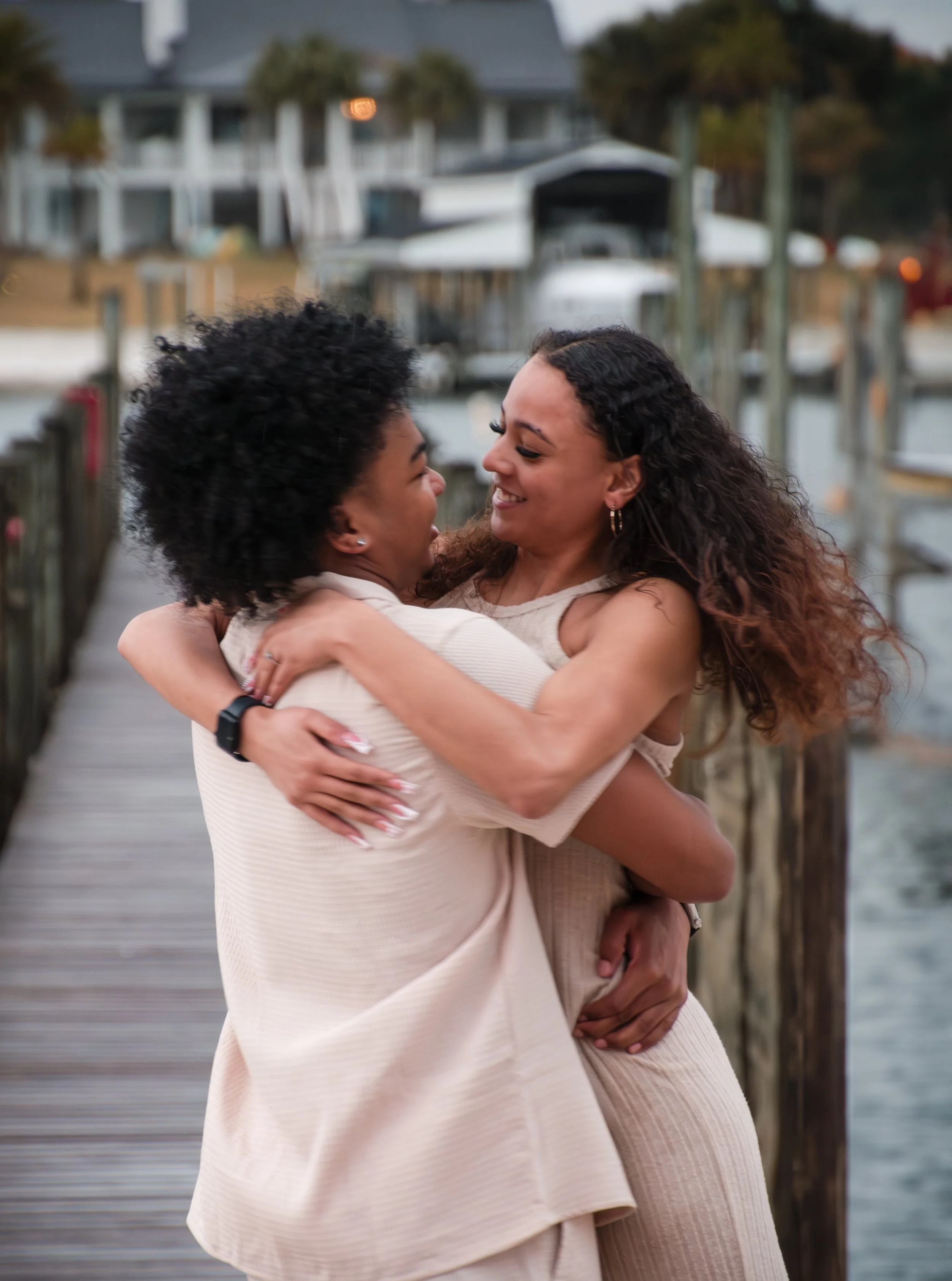 Two women are embracing and smiling at each other on a wooden dock near the water with boats and houses in the background.