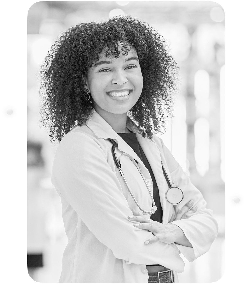 A smiling female doctor or healthcare professional with curly hair, wearing a white lab coat and stethoscope, standing confidently with arms crossed in a clinical setting.