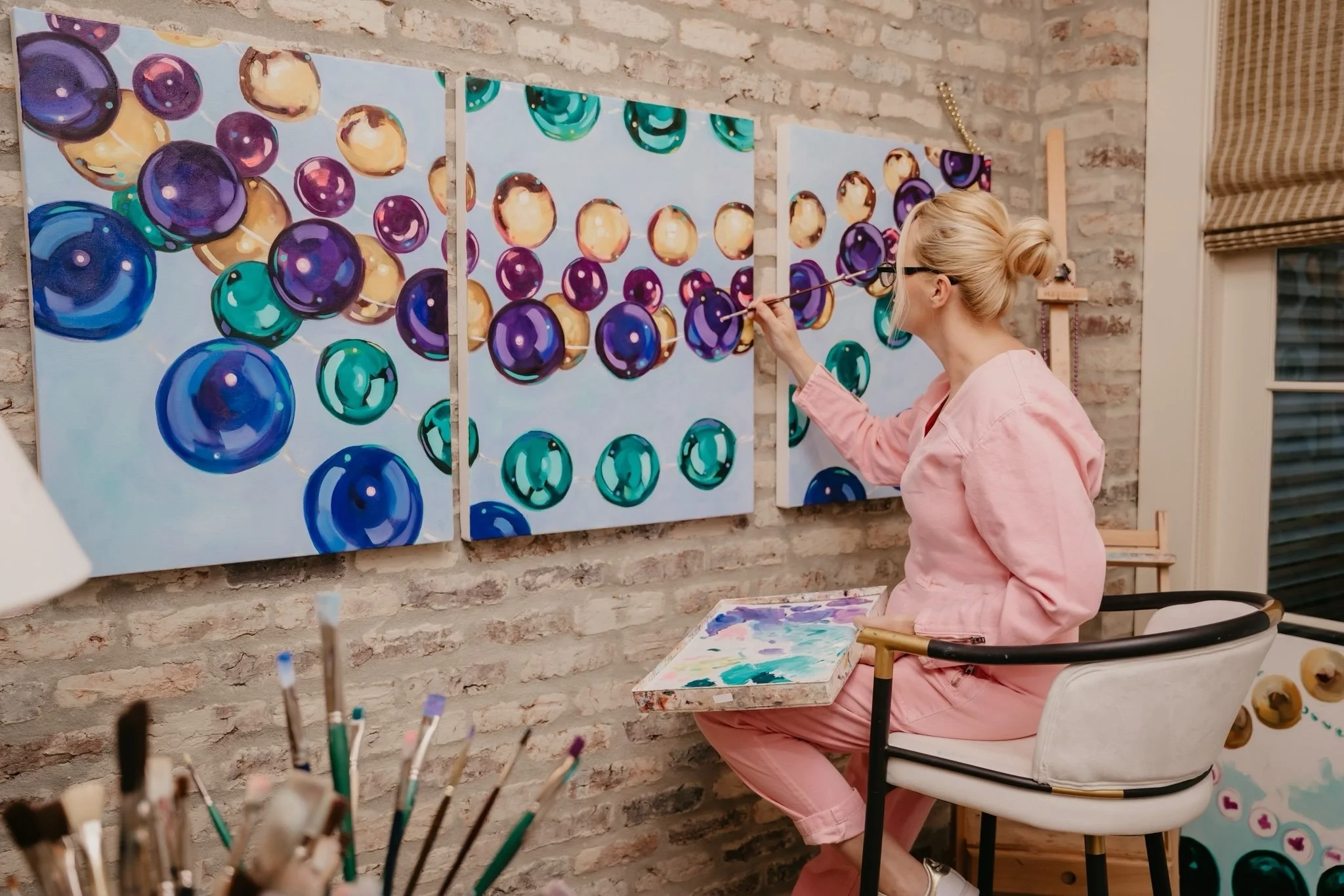 A woman painting colorful bubbles on multiple canvas panels hung on a brick wall in an art studio.