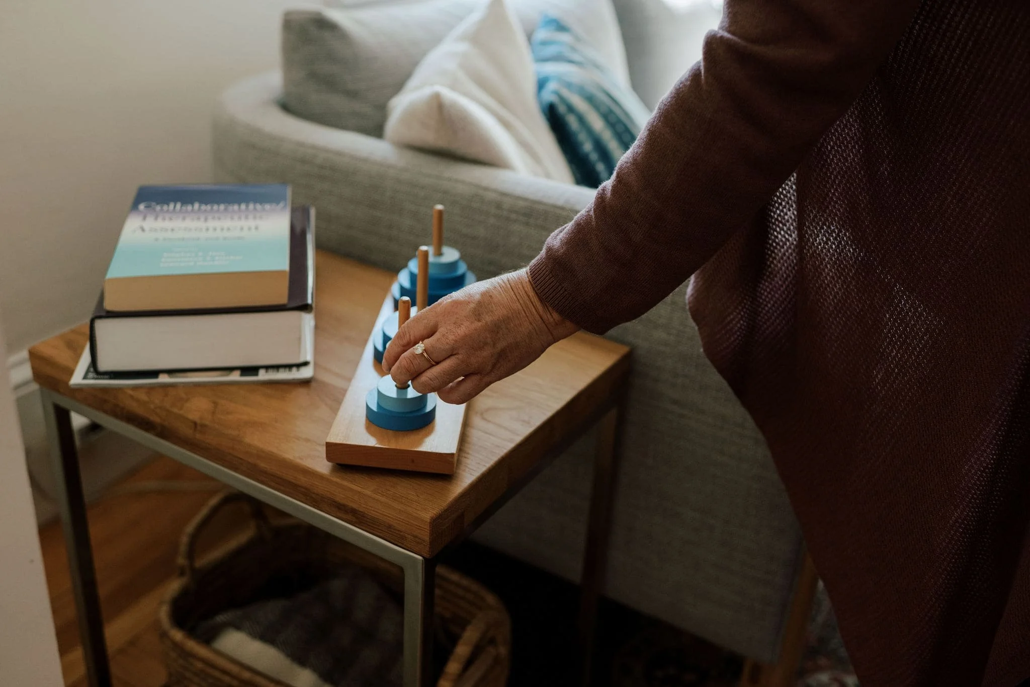 A woman with a ring on her finger adjusts a blue and white wooden toy with cylindrical pegs on a wooden board, placed on a small wooden table, near a gray sofa with pillows.