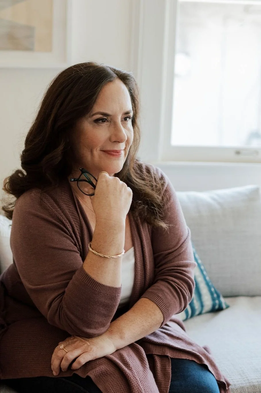 A woman with long brown hair sitting on a beige sofa near a window, holding a pair of glasses in her right hand, with a gentle smile on her face.