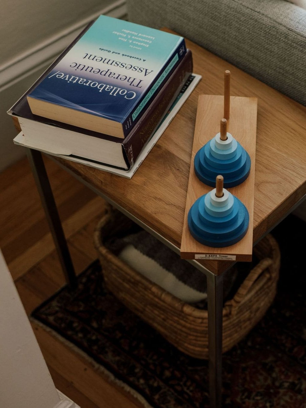Stack of two books and the set of two blue concentric ring push toys on a wooden tray on a wooden side table. A woven basket with a blanket inside is underneath the table.