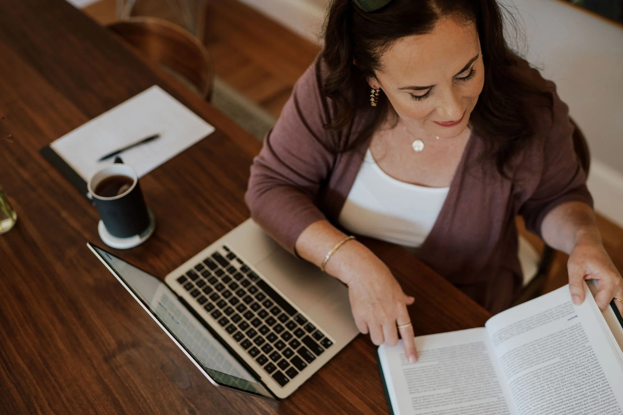 A woman sitting at a wooden table, working on a laptop, reading a book, with a cup of coffee, a notepad and pen nearby.