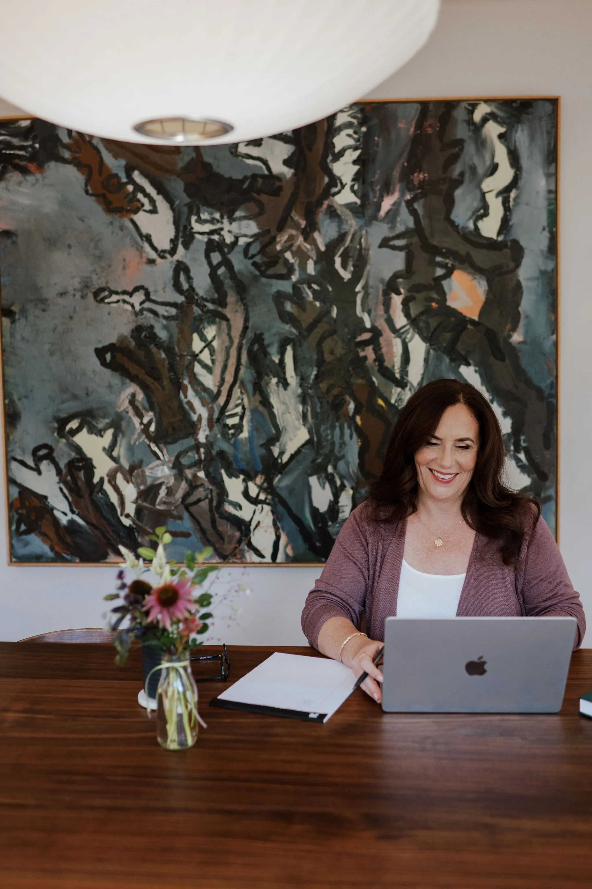 A woman sitting at a wooden table, working on a laptop, with a vase of flowers, papers, and glasses in front of her, and a large abstract painting on the wall behind her.