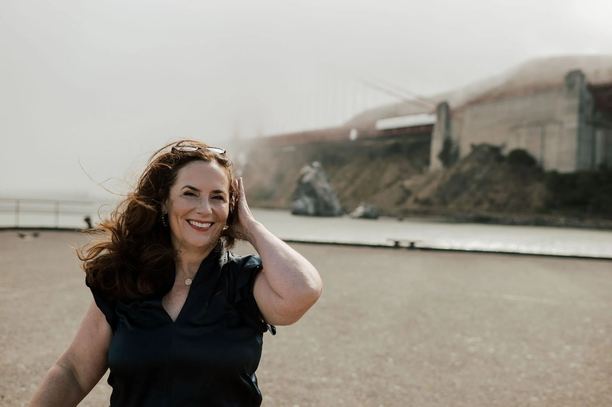 A woman with shoulder-length brown hair, smiling, holding her hair back with one hand, standing on a beach with a foggy bridge and rocky cliffs in the background.