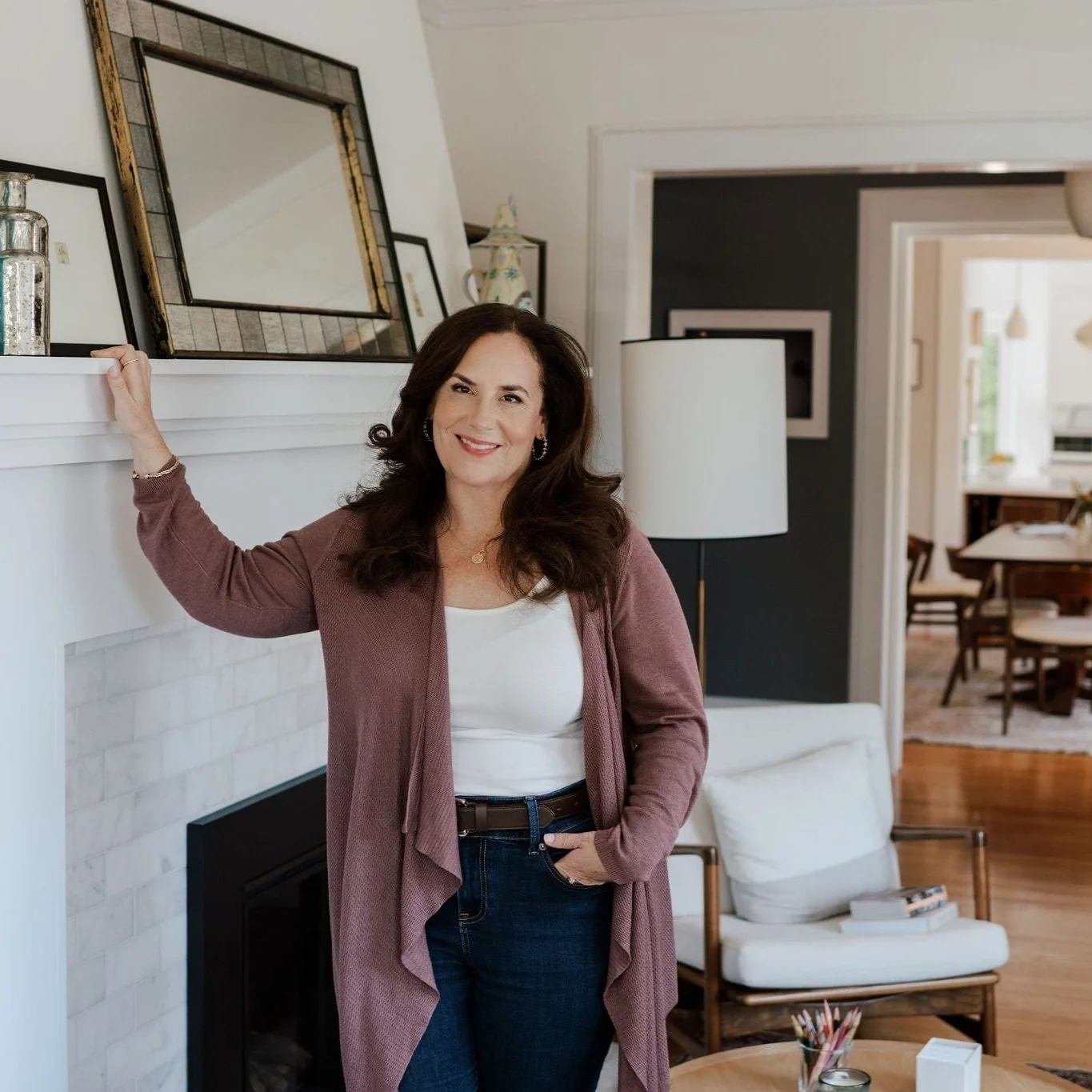 A woman with long dark hair wearing a white top, pink cardigan, and dark jeans is standing near a fireplace with her left hand resting on the mantle. She is smiling in a living room with a mirror, framed artwork, a lamp, a white armchair, and a wooden dining area visible in the background.