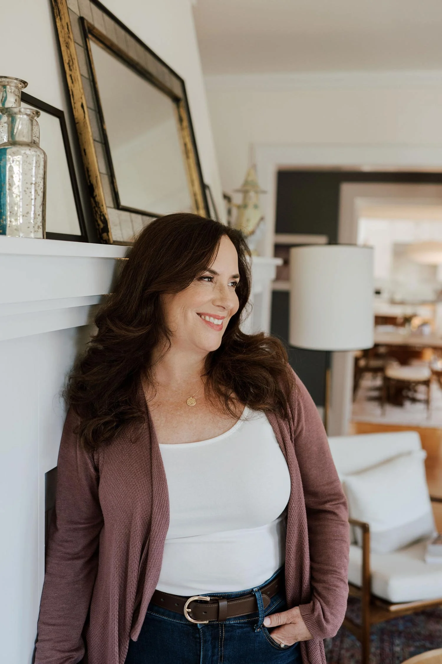 A smiling woman with dark brown hair, wearing a white top, a pinkish cardigan, blue jeans, and a brown belt, leaning against a white wall in a cozy home interior.