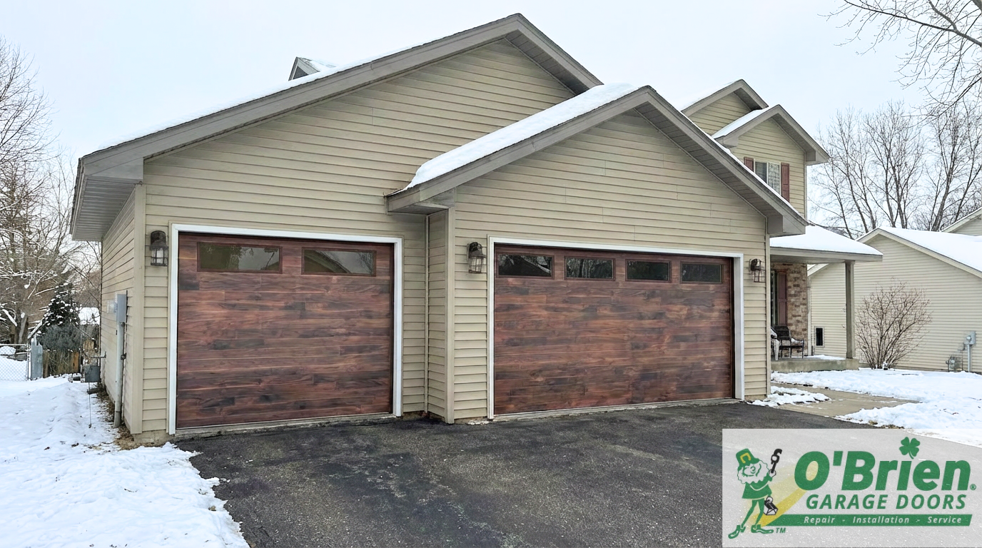 A two-car garage with tan siding and wooden garage doors, snow on the roof and ground, and a neighboring house in the background, with the O'Brien Garage Doors logo in the bottom right corner.