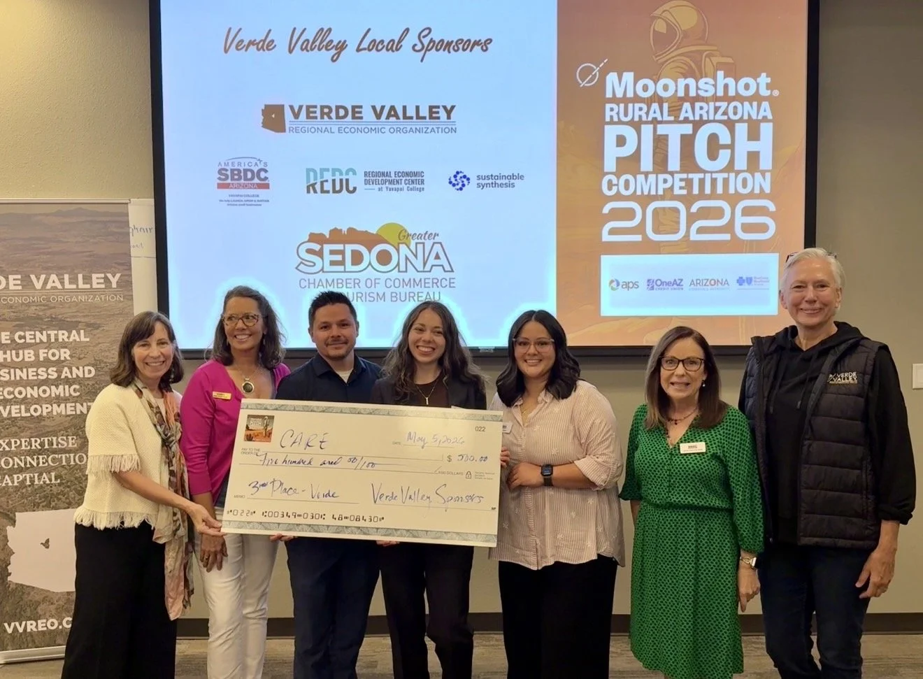 Two people holding a large check at a presentation event for the Moonshot Arizona Pitch Competition 2023, with sponsors displayed on a screen in the background.