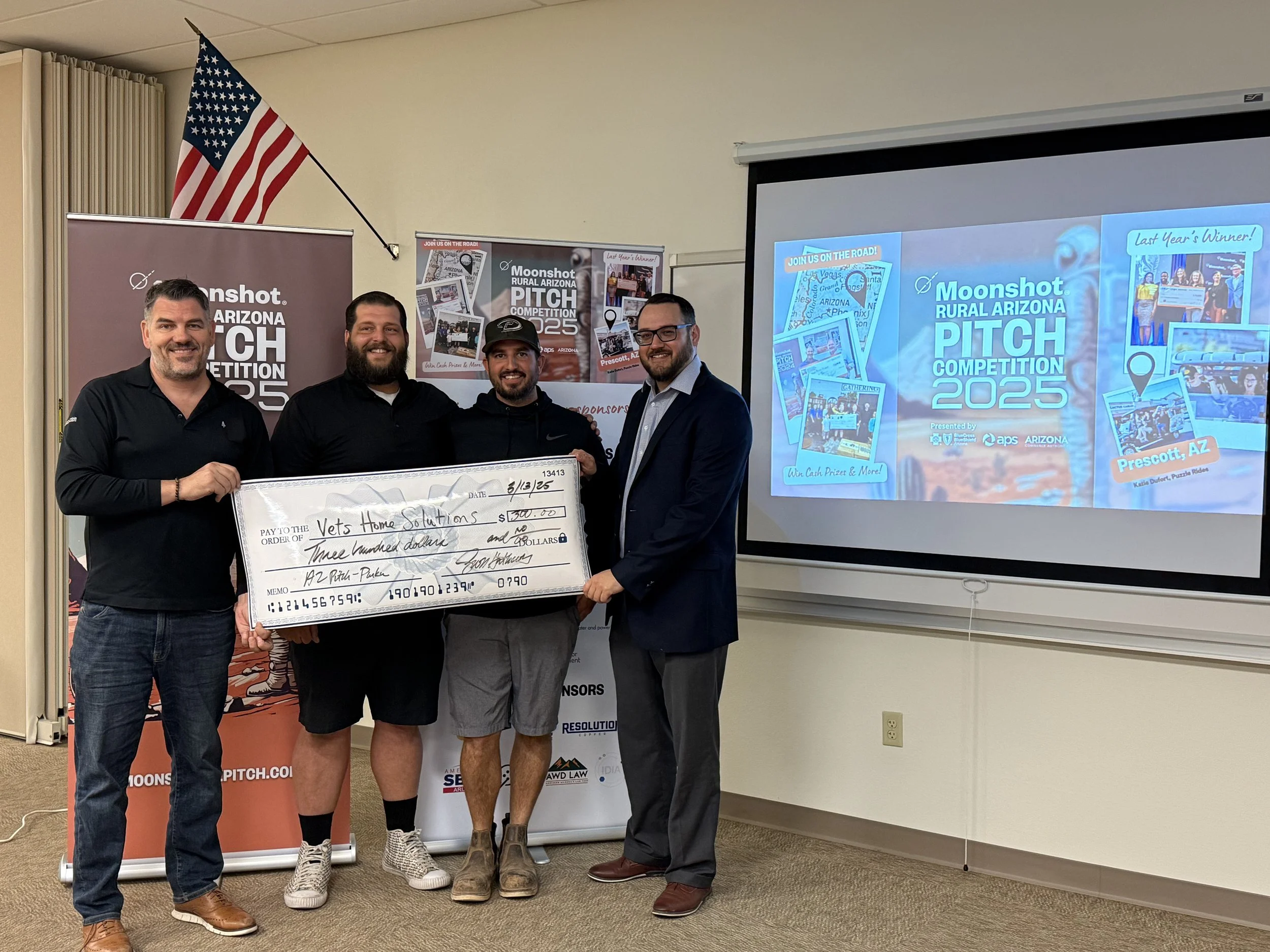 Four people standing, holding a large check for "Vets Home Solutions" from the Moonshot Rural Arizona Pitch Competition 2025 event. An American flag is displayed behind them, along with banners and a presentation screen.