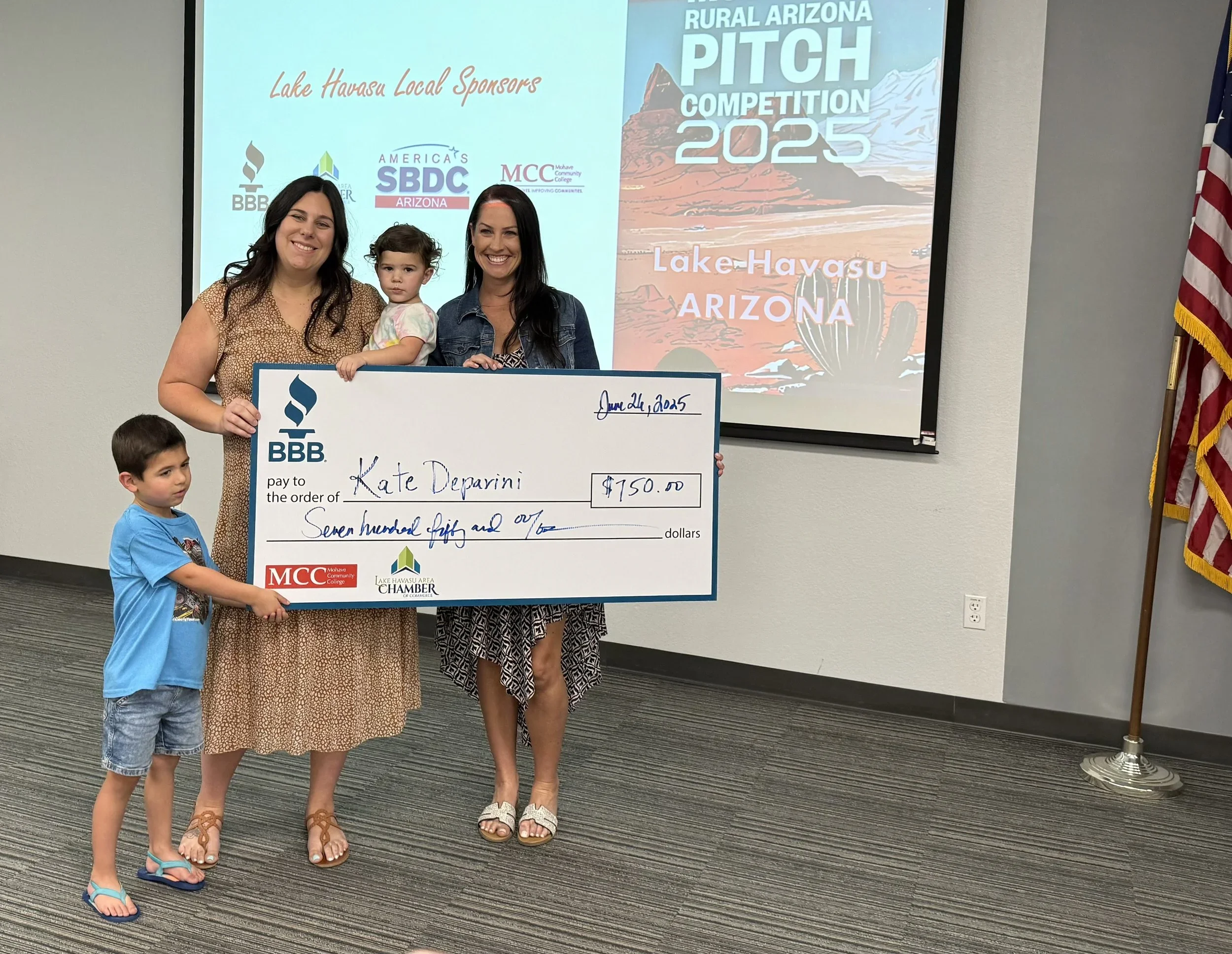 Group of three women and two children holding a large check during a ceremony, with a presentation screen in the background displaying 'Rural Arizona Pitch Competition 2025' and 'Lake Havasu Arizona.' The women are smiling, and the children are standing close, one of whom is in the women's arms. The check is made out to Kate Deparini for $150, with logos for BBB, MCC, and Lake Havasu Chamber.