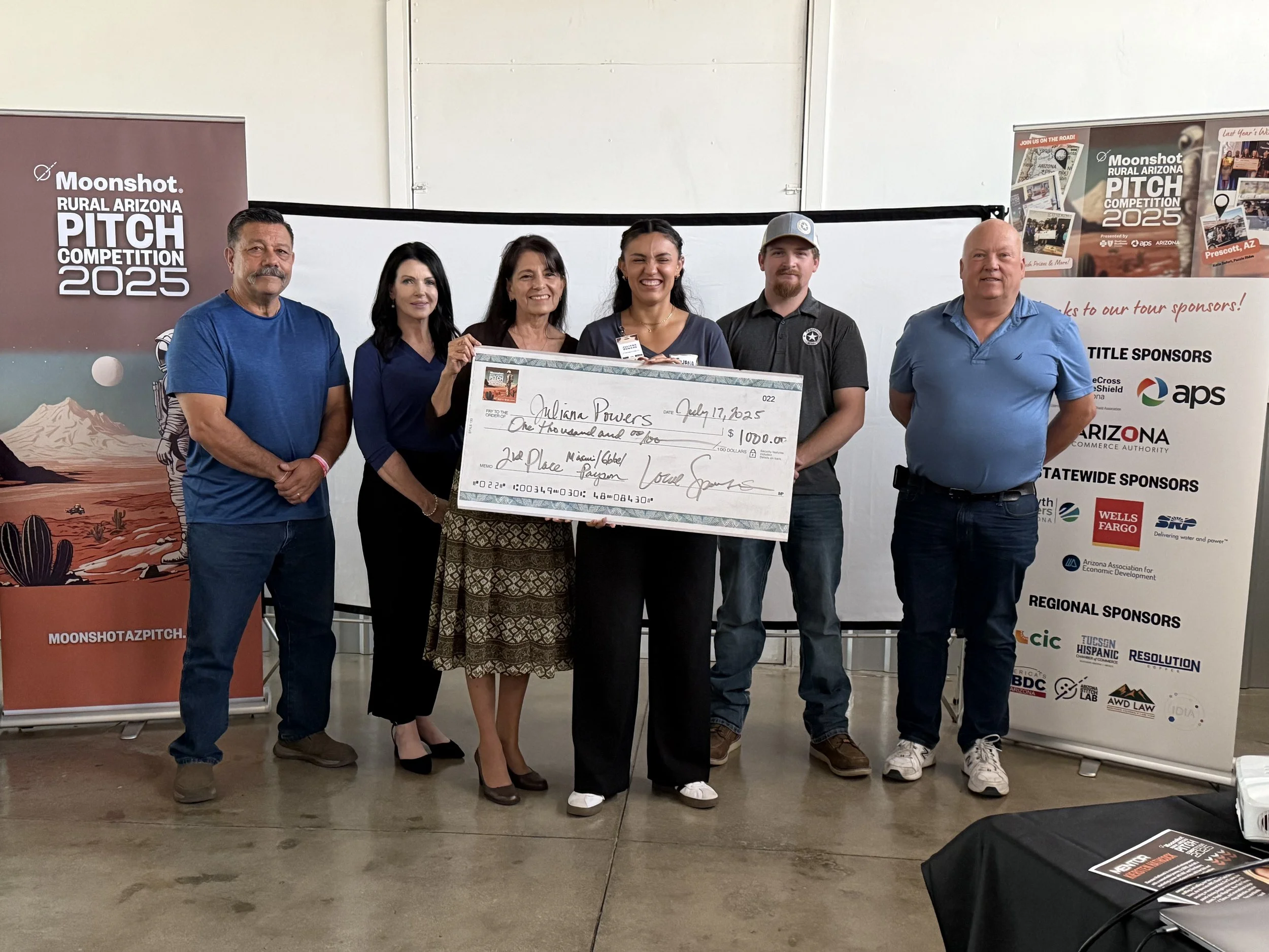 Group of six people standing indoors holding a large check for $1,000, celebrating an award at the Moonshot Rural Arizona Pitch Competition 2025, with banners and sponsor signs in the background.