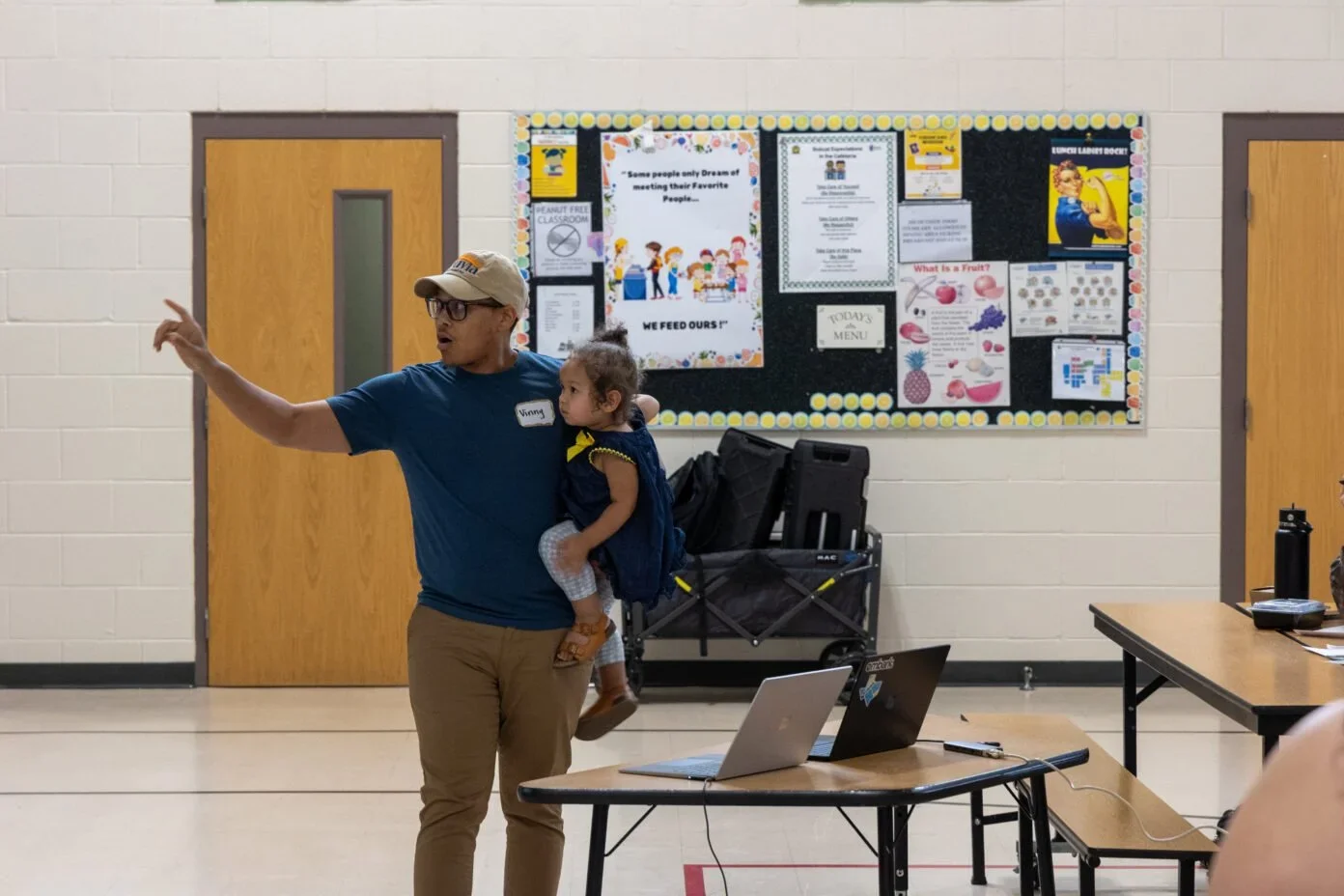 A man wearing glasses and a beige cap, with a name tag that says 'Vinny,' holds a young girl in his arm, pointing to the side while giving a presentation or speech in a room with educational posters on bulletin boards.